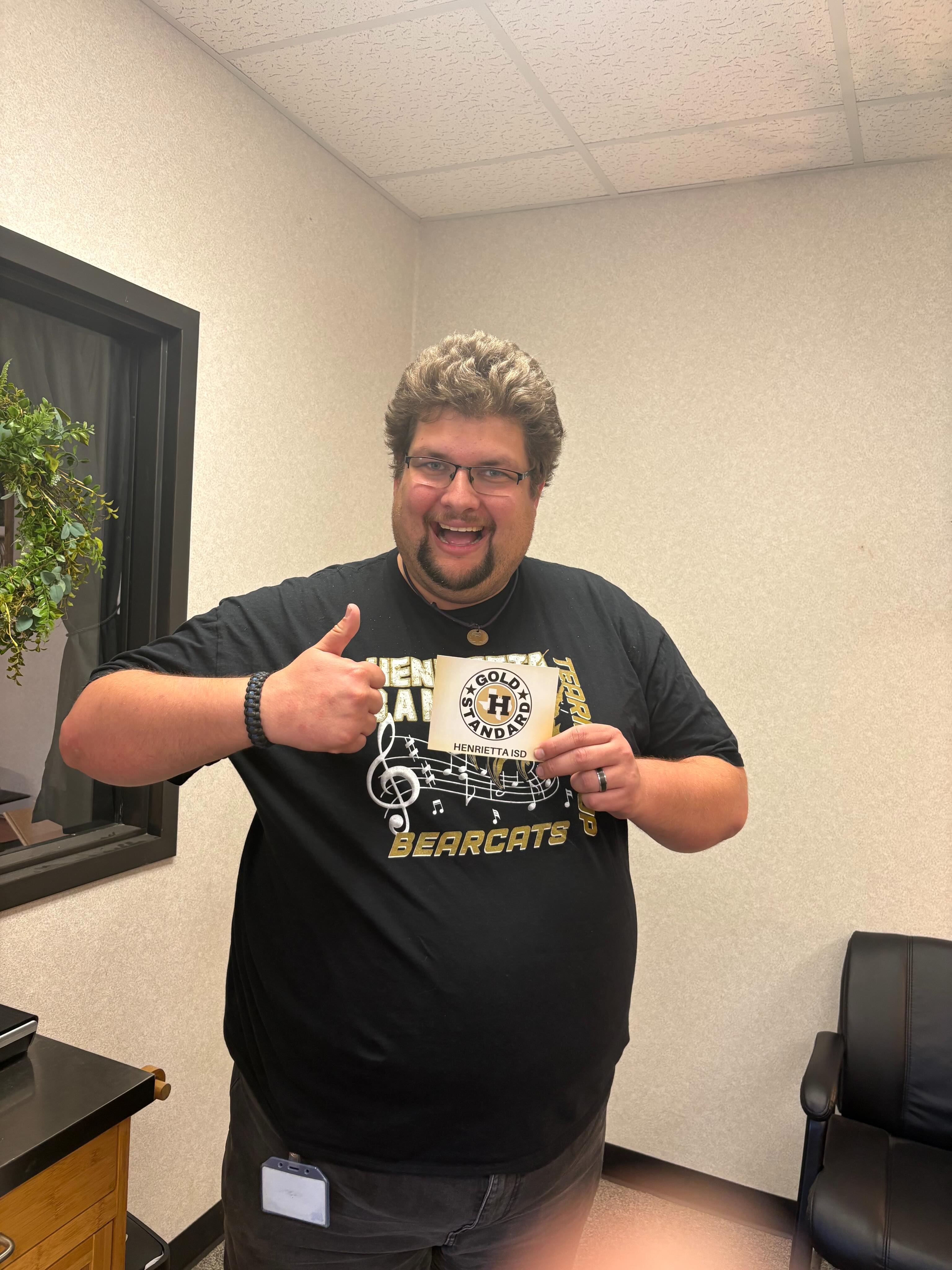 Person in a black t-shirt with "Bearcats" and a music note standing in a room with a plant.