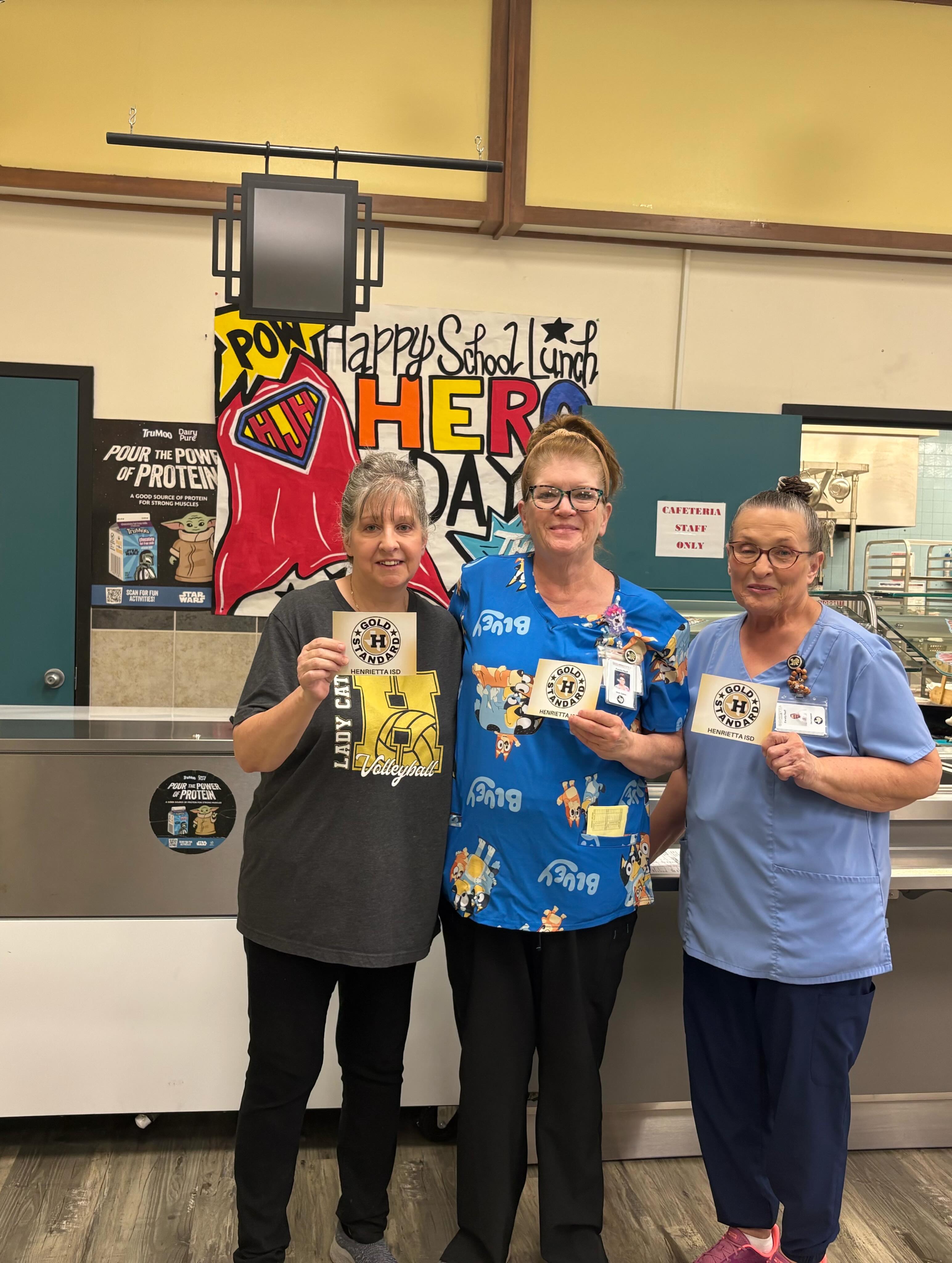 Three women stand together in a lobby, holding cards. One woman wears a blue shirt, another a gray shirt, and the third wears blue scrubs. Behind them, a banner says "Happy School Year."