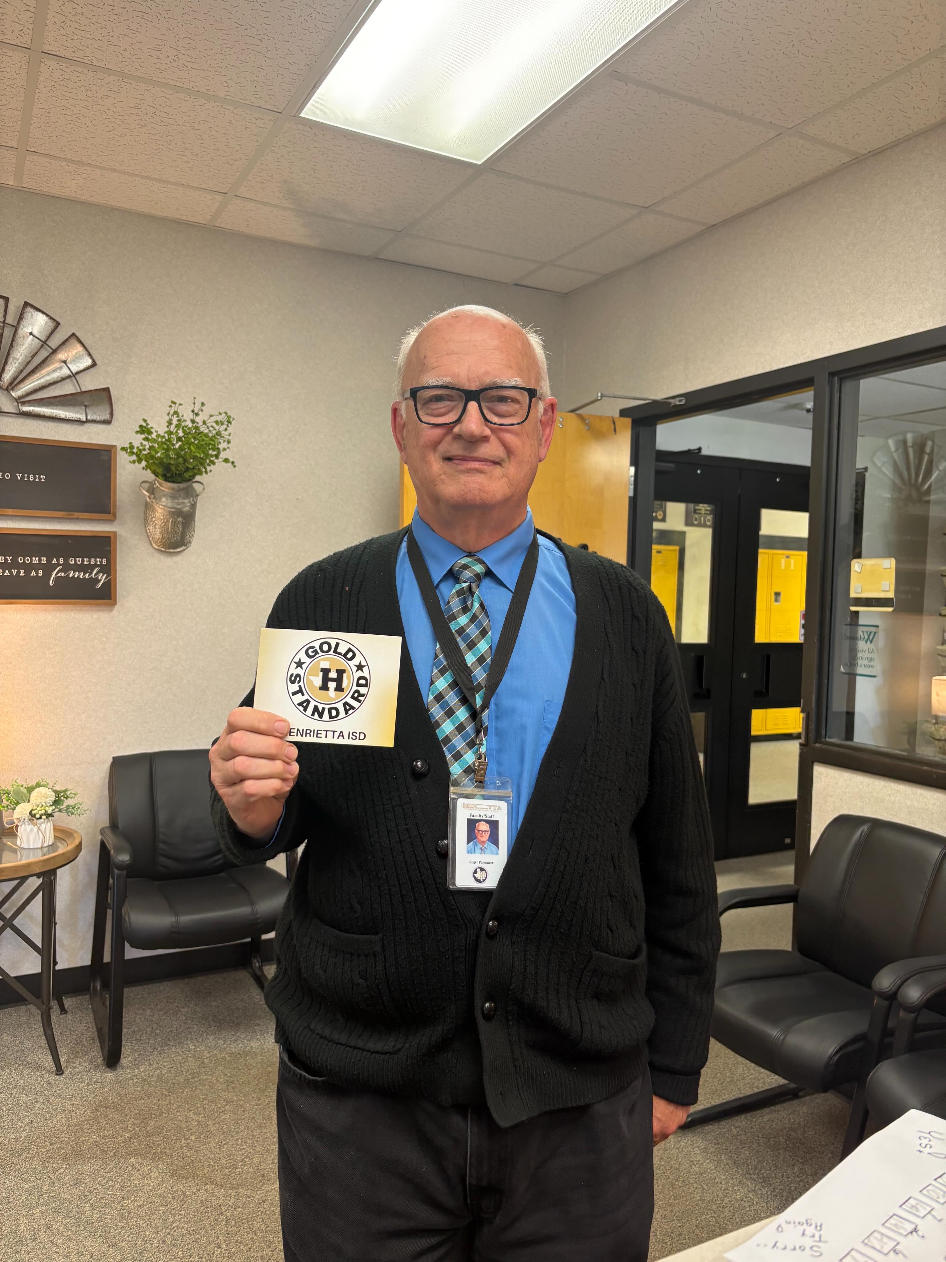 Man wearing glasses, a blue shirt, and a black cardigan holds a card with a logo.
