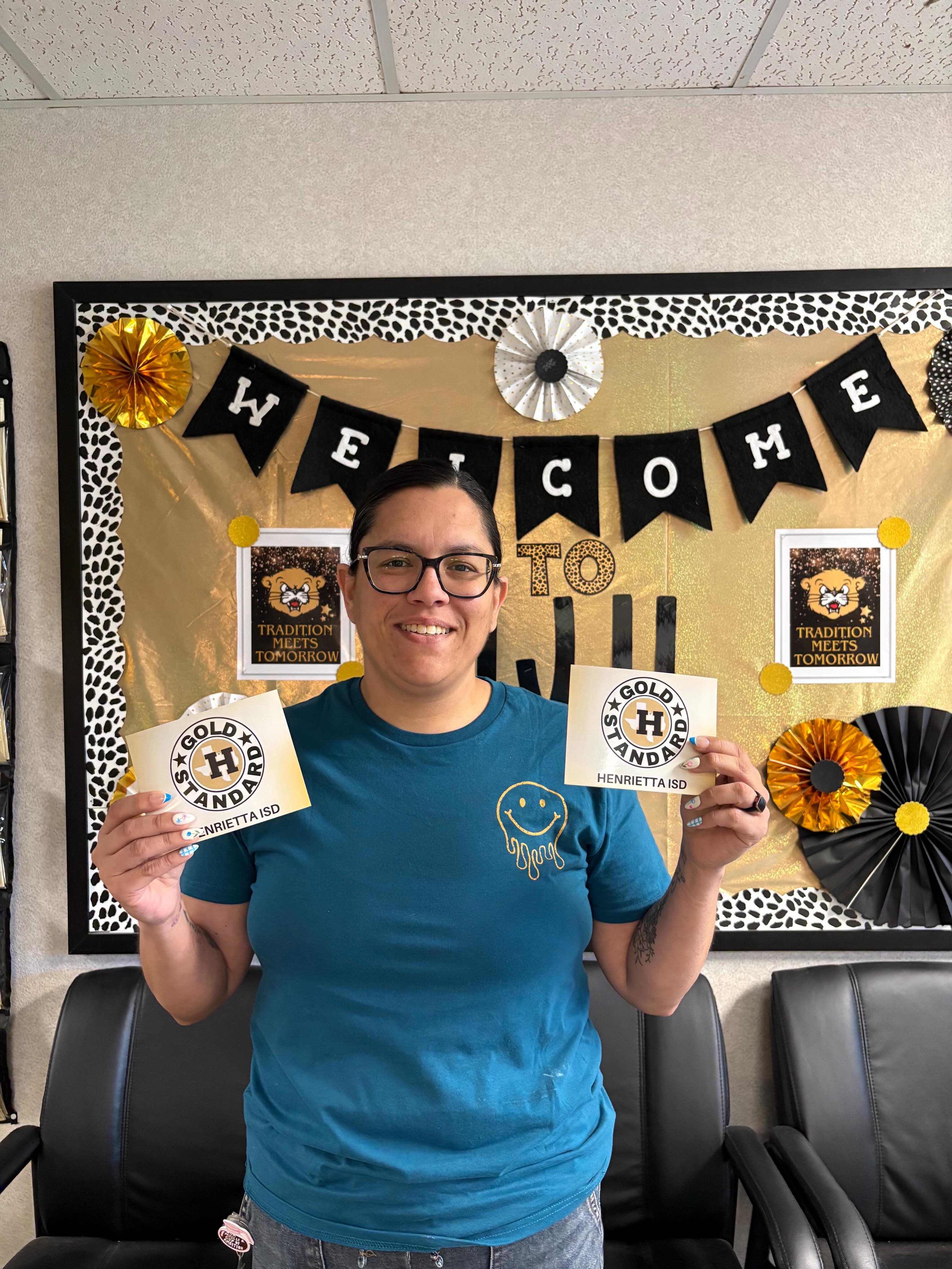 A woman in a blue shirt holds two cards with 'GOLDEN HANDS' in front of a bulletin board with a 'WELCOME' banner and black chairs.
