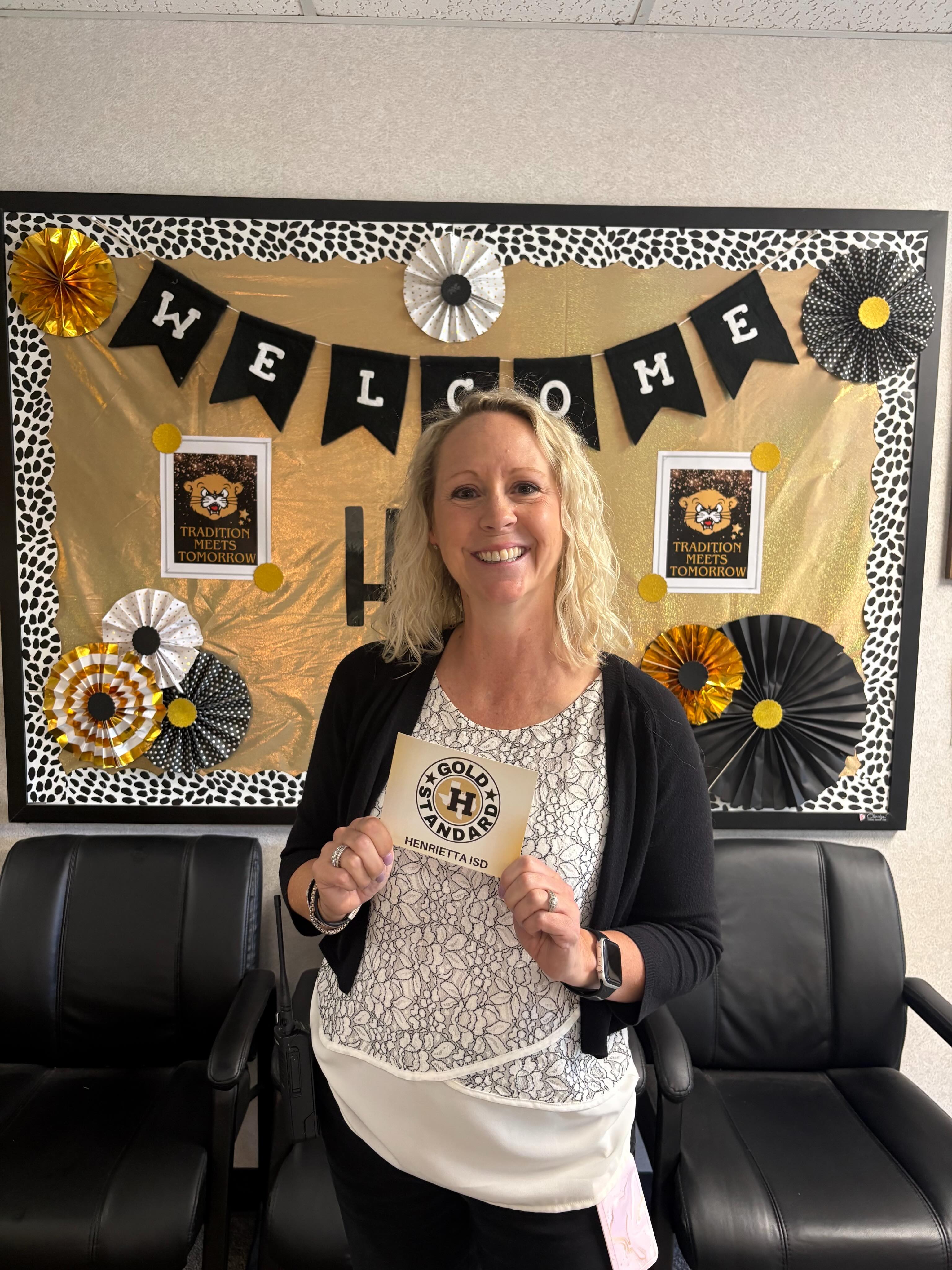 Woman standing in a room with black chairs, holding a paper award. A decorative bulletin board with a "Welcome" banner is behind her.