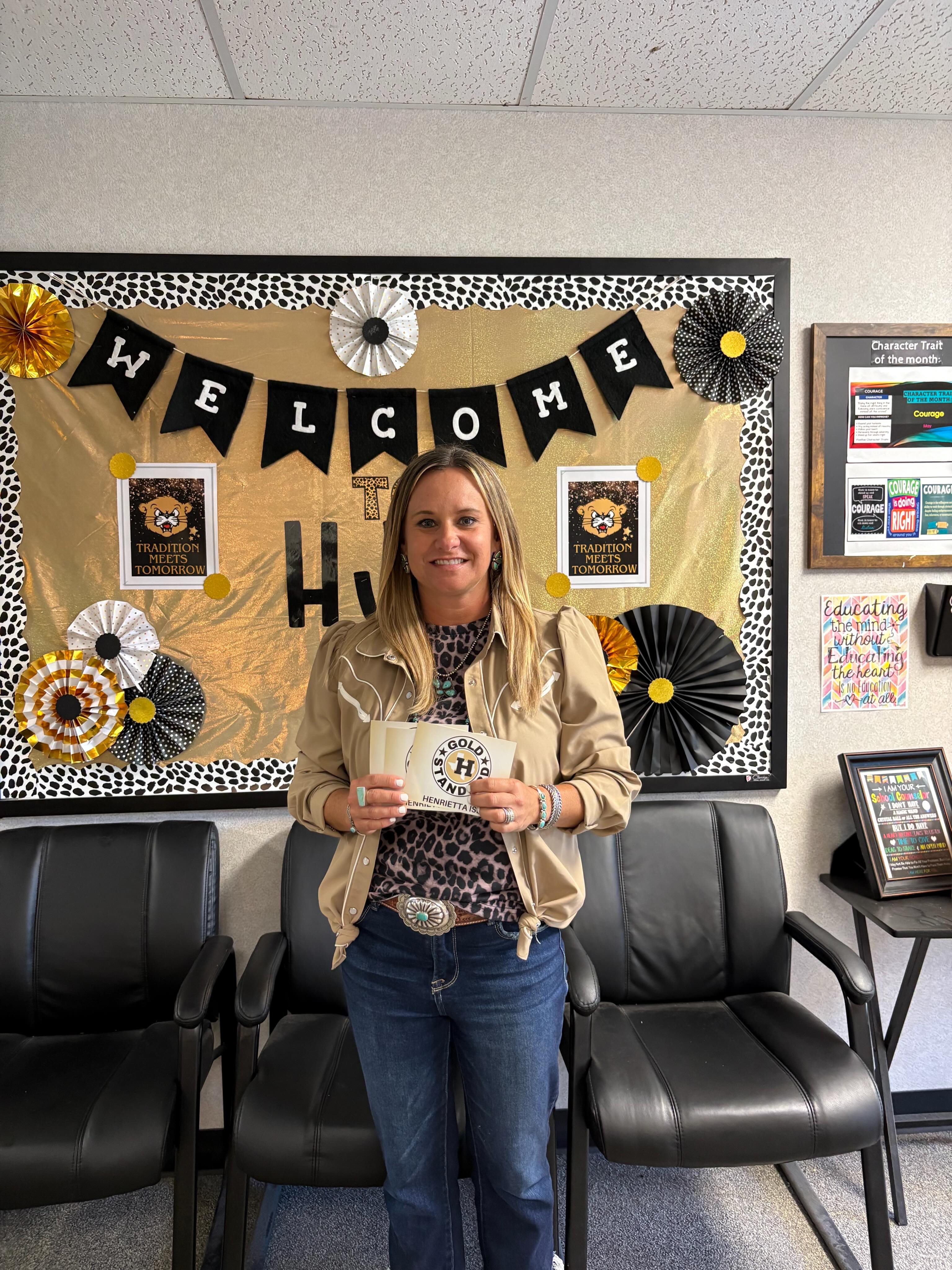 Woman holding a check in a room with black chairs, welcome banner, and a framed picture.