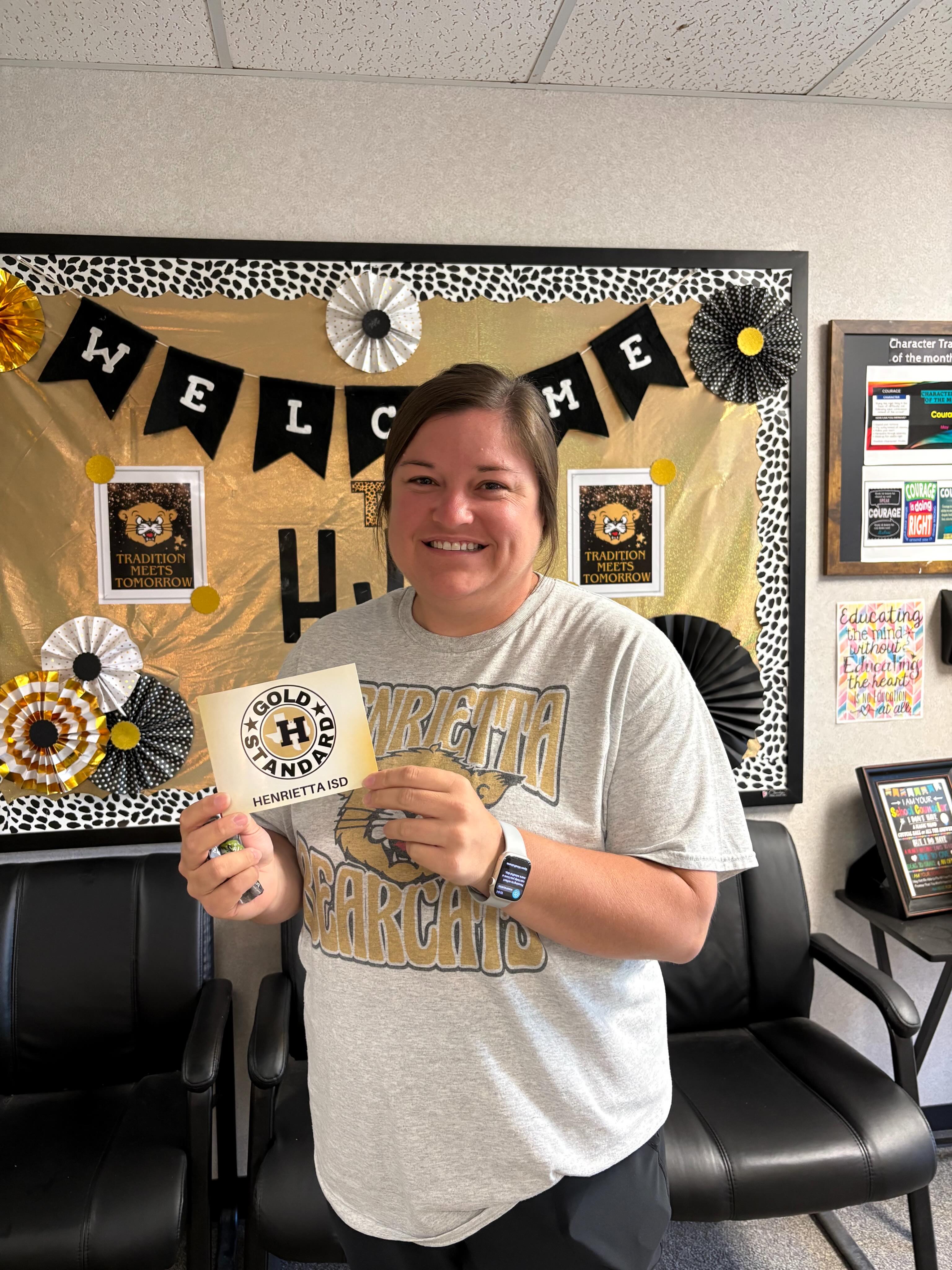 Woman holding certificate in front of bulletin board with black and yellow decorations and text "Welcome".