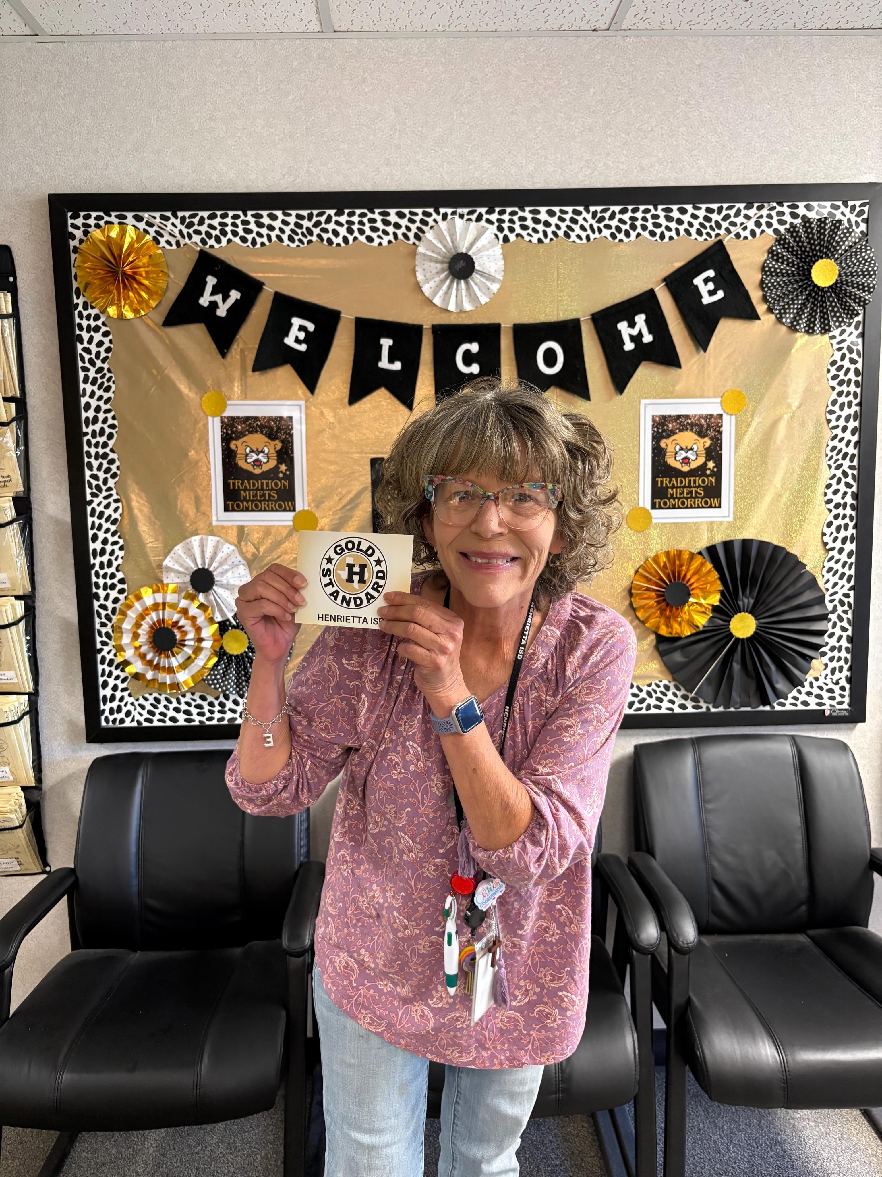 An older woman holds a small card in front of her, smiling. Behind her is a colorful bulletin board with "WELCOME" written in large letters.
