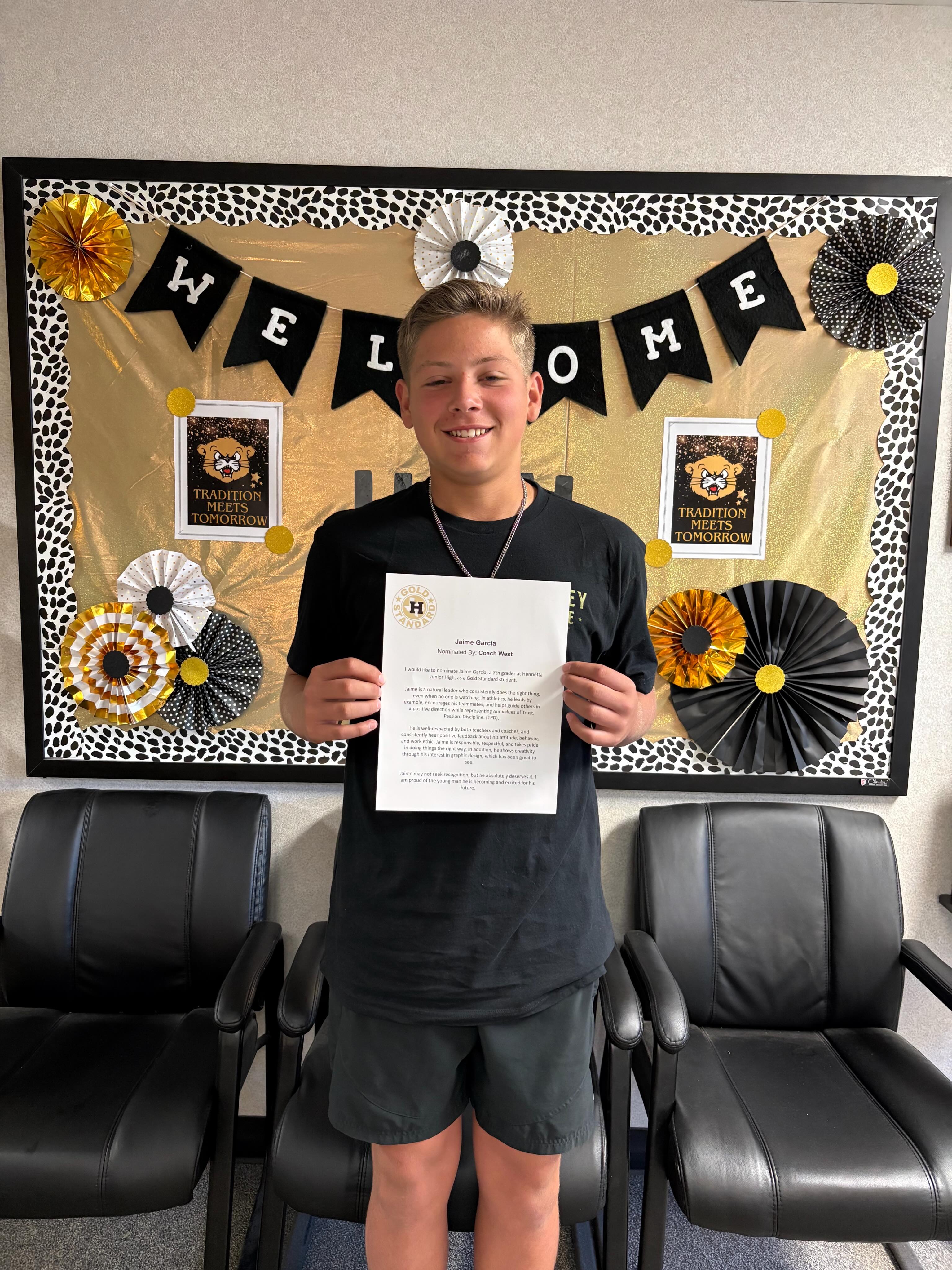 A young person stands holding a certificate in front of a bulletin board with a "Welcome" banner.