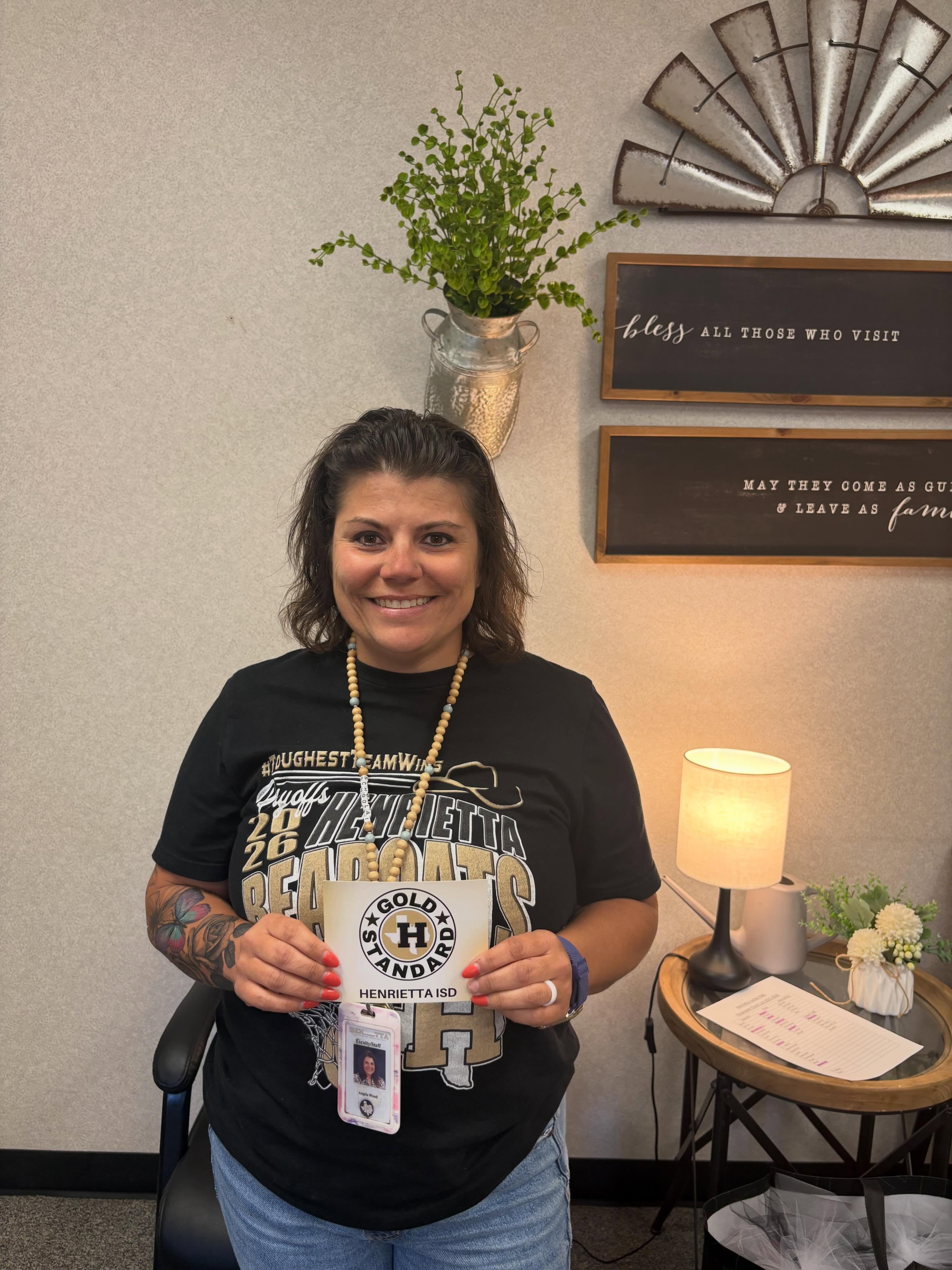 Woman in black t-shirt holds an ID card. Behind her, decorative items on a wall and table.