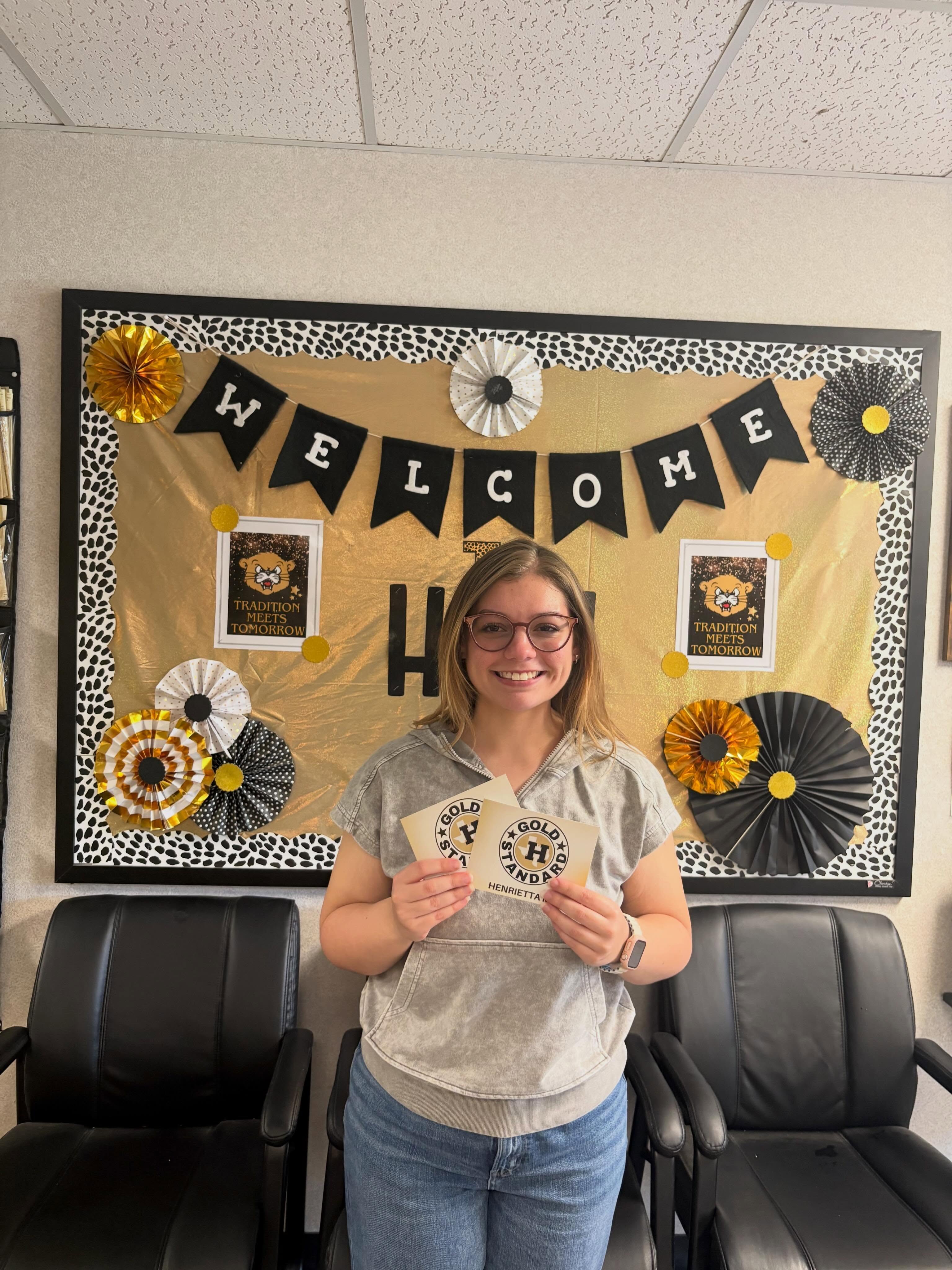 A woman in a gray shirt and glasses stands in a room holding a certificate. Behind her, a bulletin board has a "Welcome" banner.
