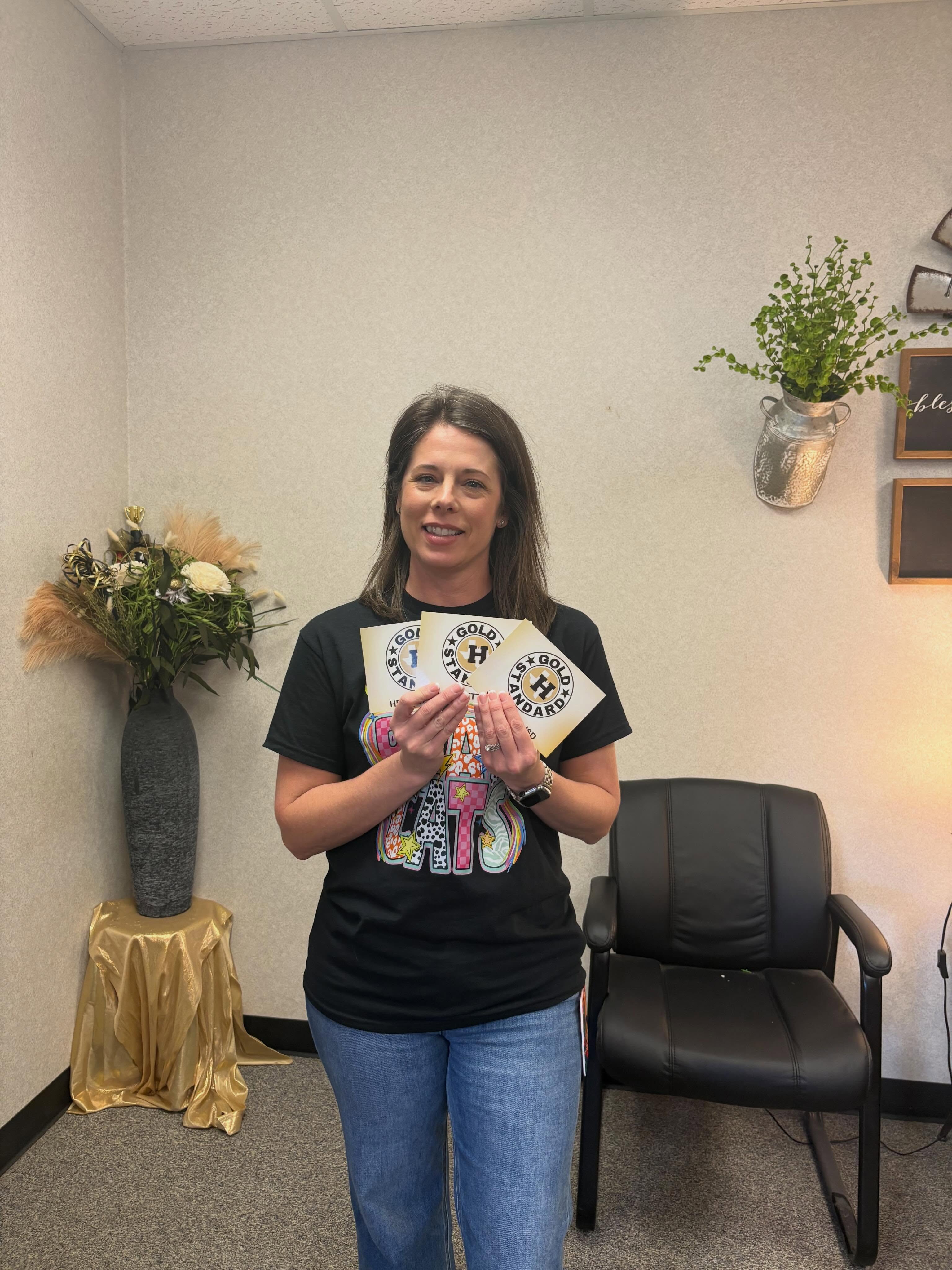 Woman holding cards and smiling in a room with a flower vase and a black chair.