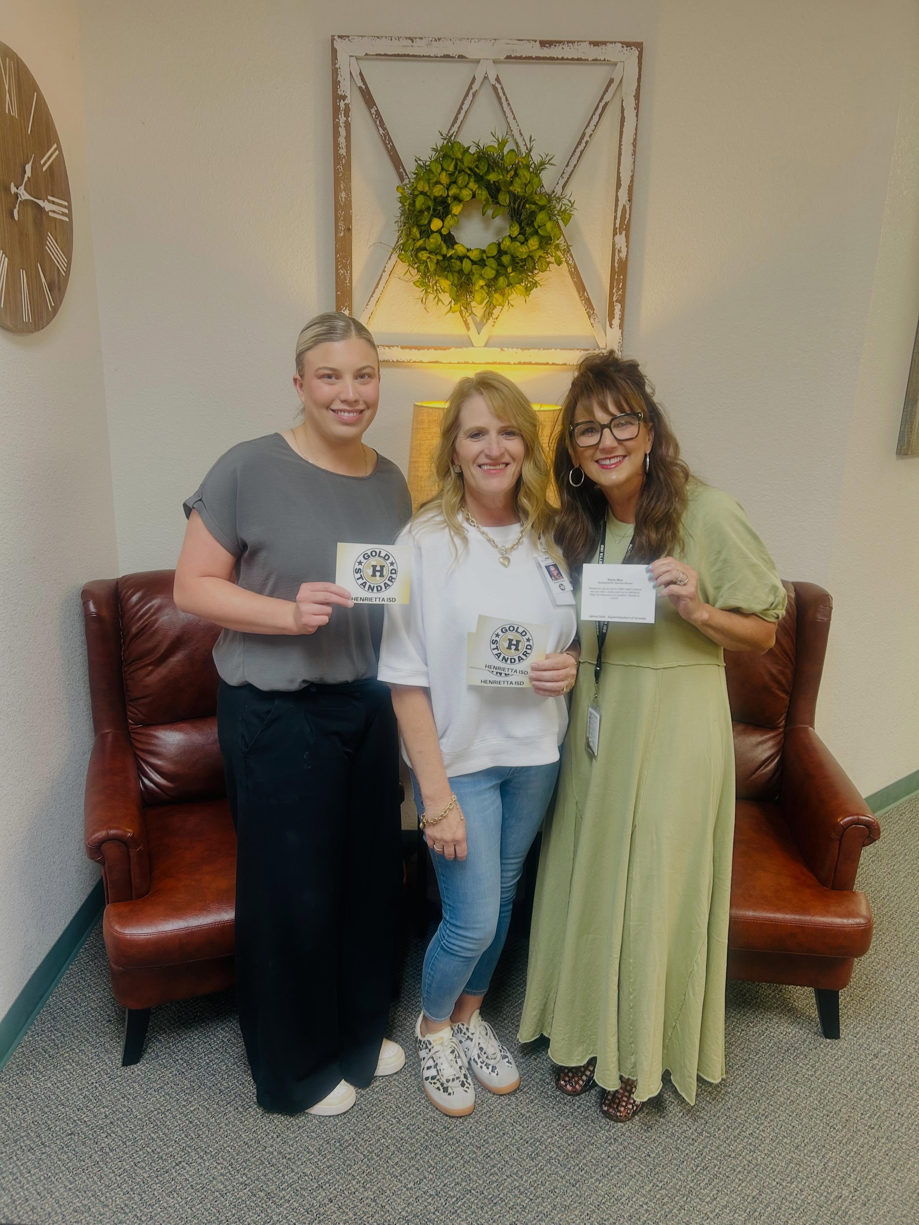 Three women stand in a room, holding certificates. One wears a green dress, another a gray shirt, and the third wears black pants. Behind them are two armchairs, a wreath, and a wall clock.