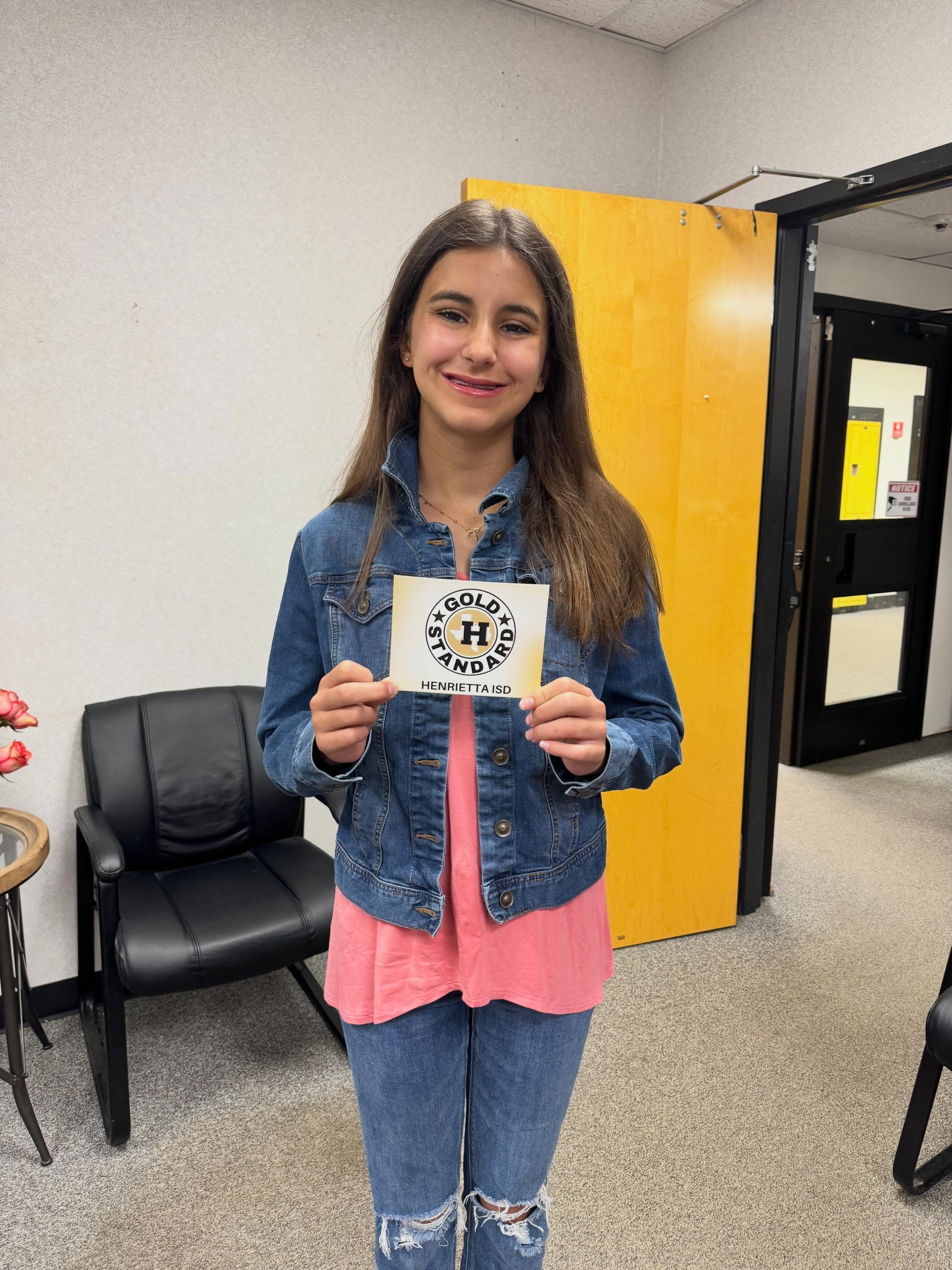 A young girl in a pink shirt and denim jacket holds a rectangular sign. She is standing in a room with a yellow door and black chair.
