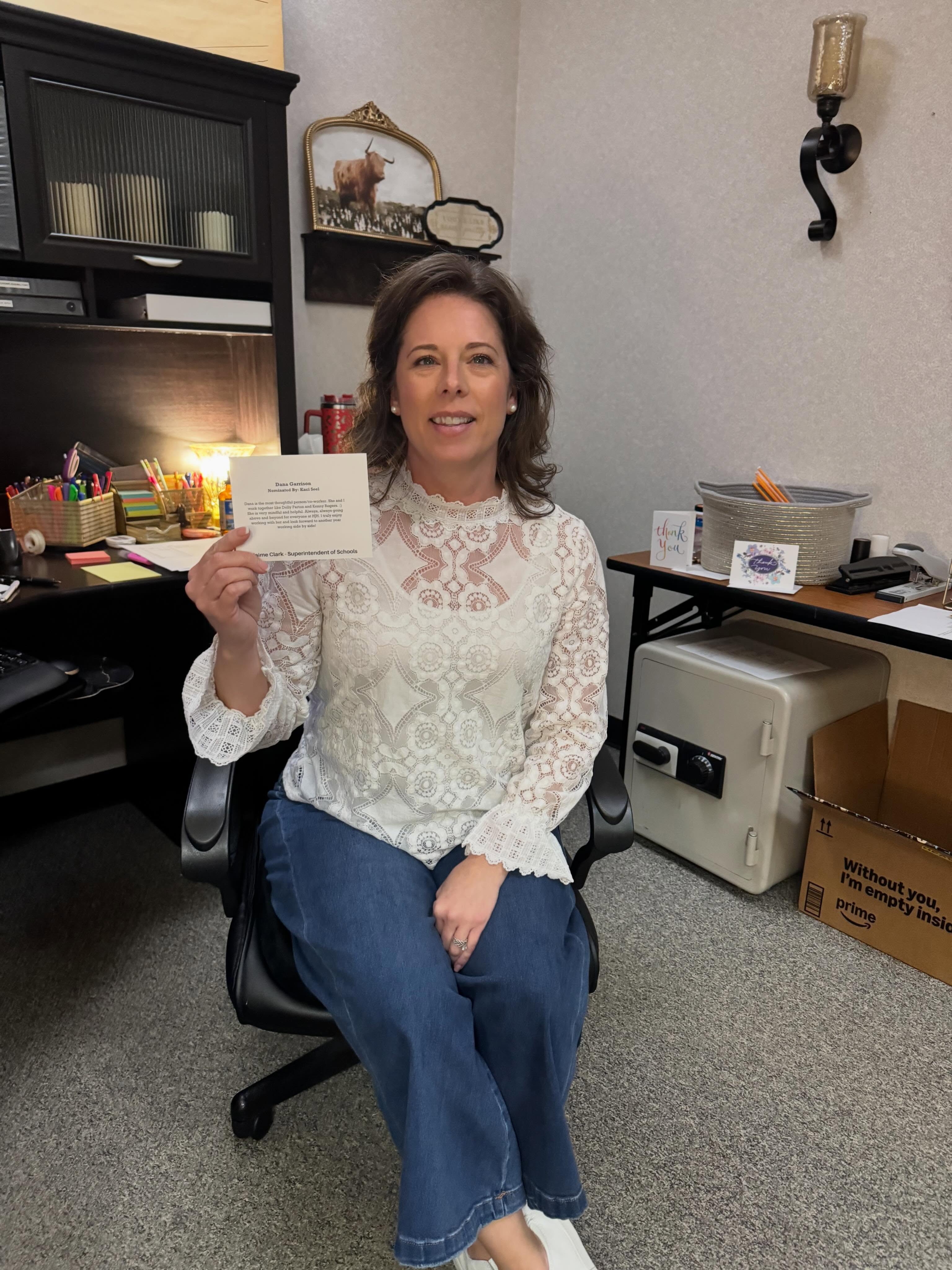 A woman in a white lace blouse and blue jeans sits in a chair holding an envelope. Behind her is a desk and a small table.