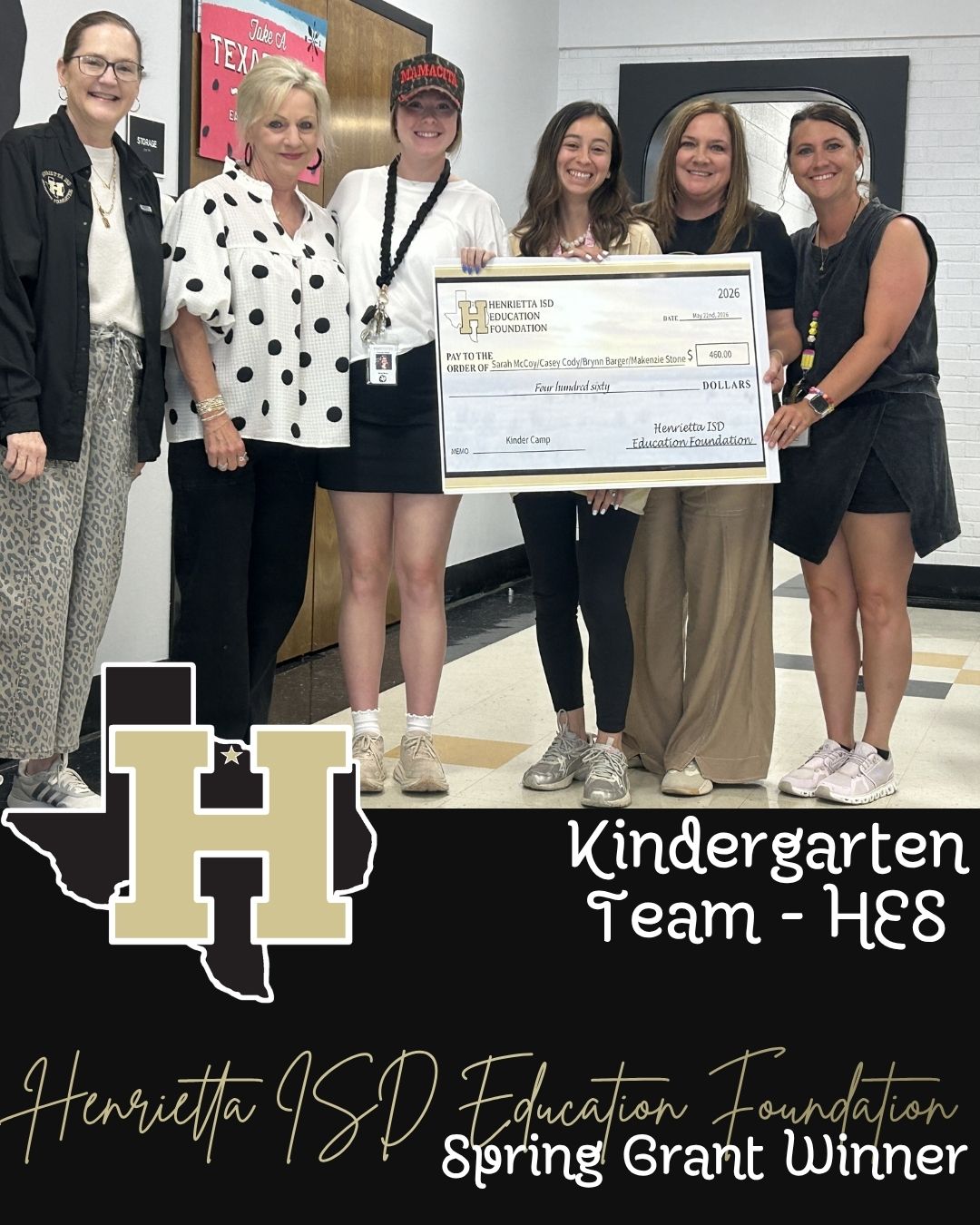 Six women, five in shorts, hold a check and stand near a school logo. They are smiling.
