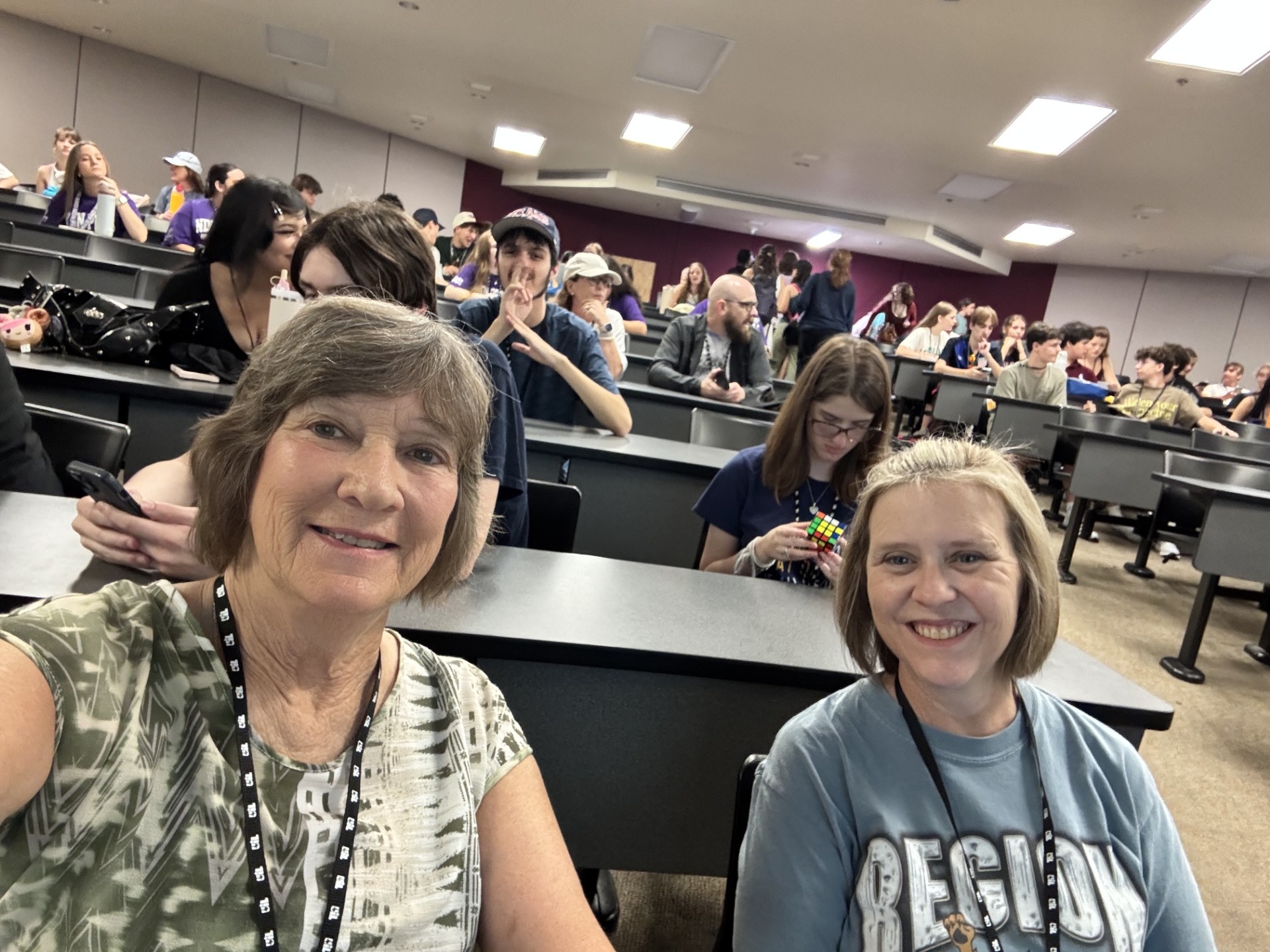 Two smiling women seated in a classroom with rows of desks and people in the background.