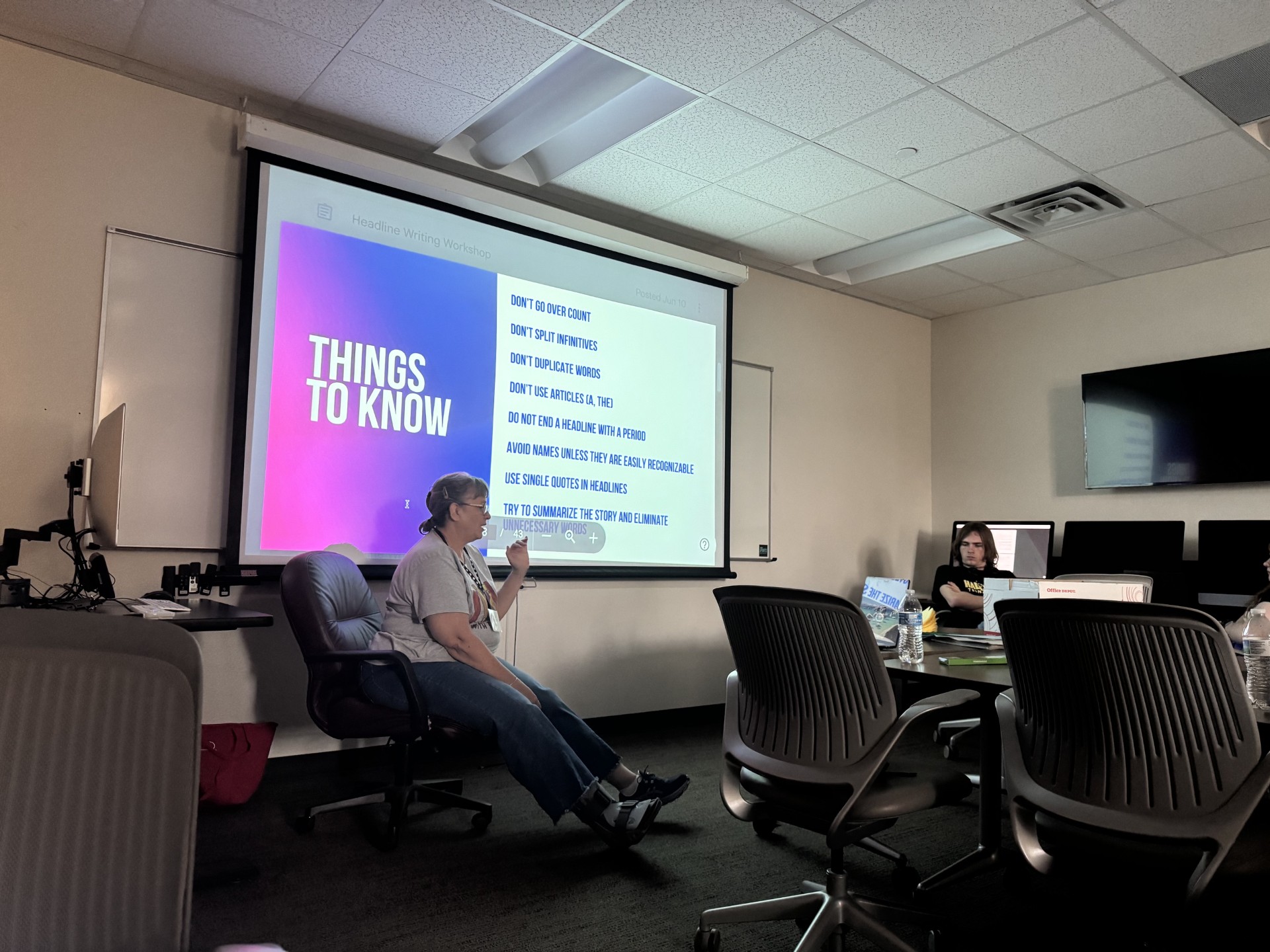 Two people in a meeting room. One is seated, holding a microphone. A large projector screen shows "THINGS TO KNOW."