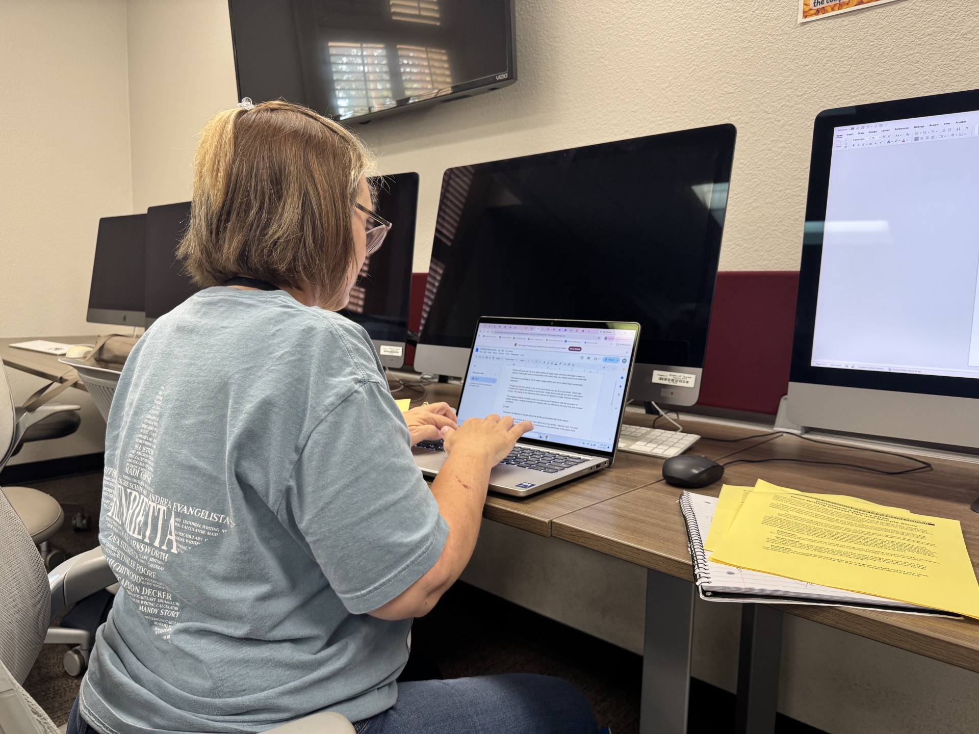 A woman with glasses sits at a desk with several monitors, typing on a laptop.