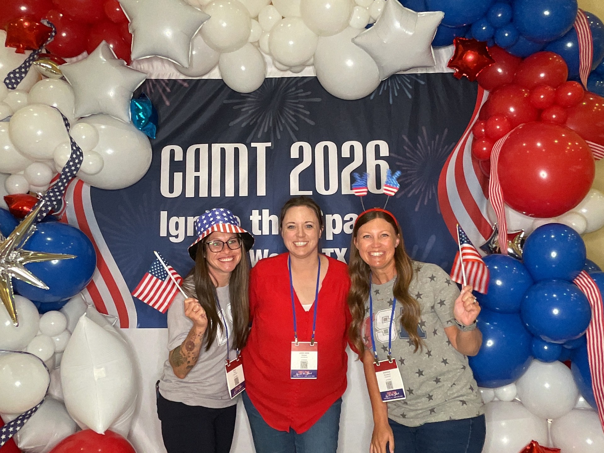 Three women pose in front of a large balloon backdrop with an American flag banner reading "CAMT 2026."