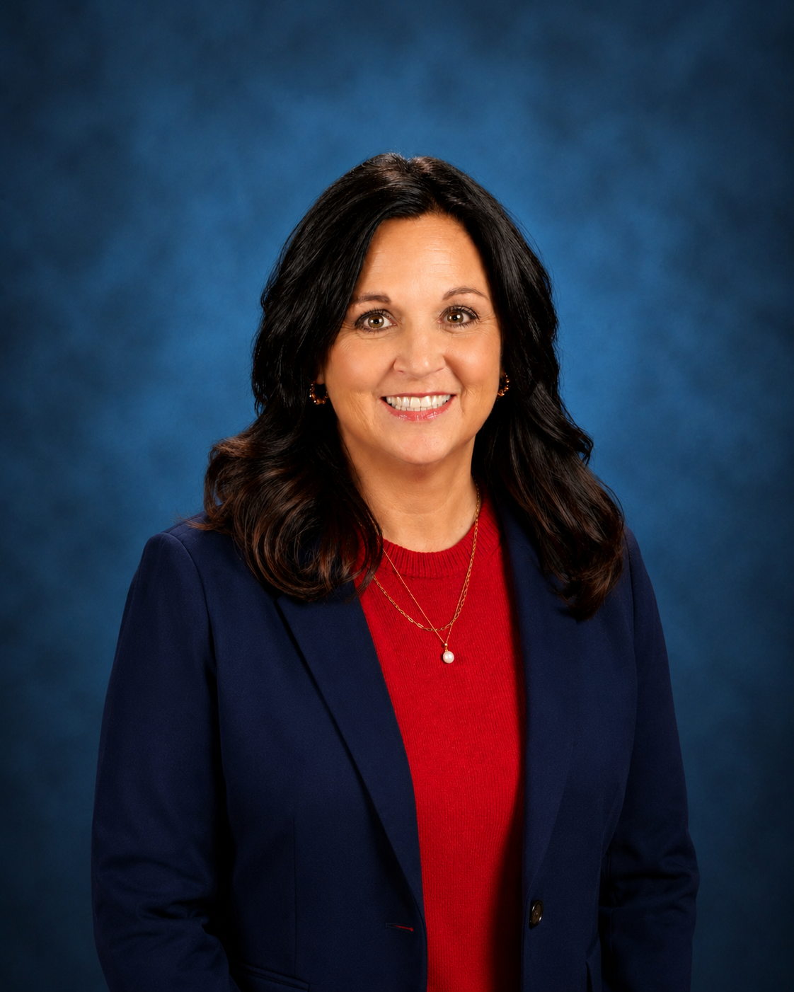 Woman with dark wavy hair wearing a red top, blue blazer, and a necklace with a small pendant.