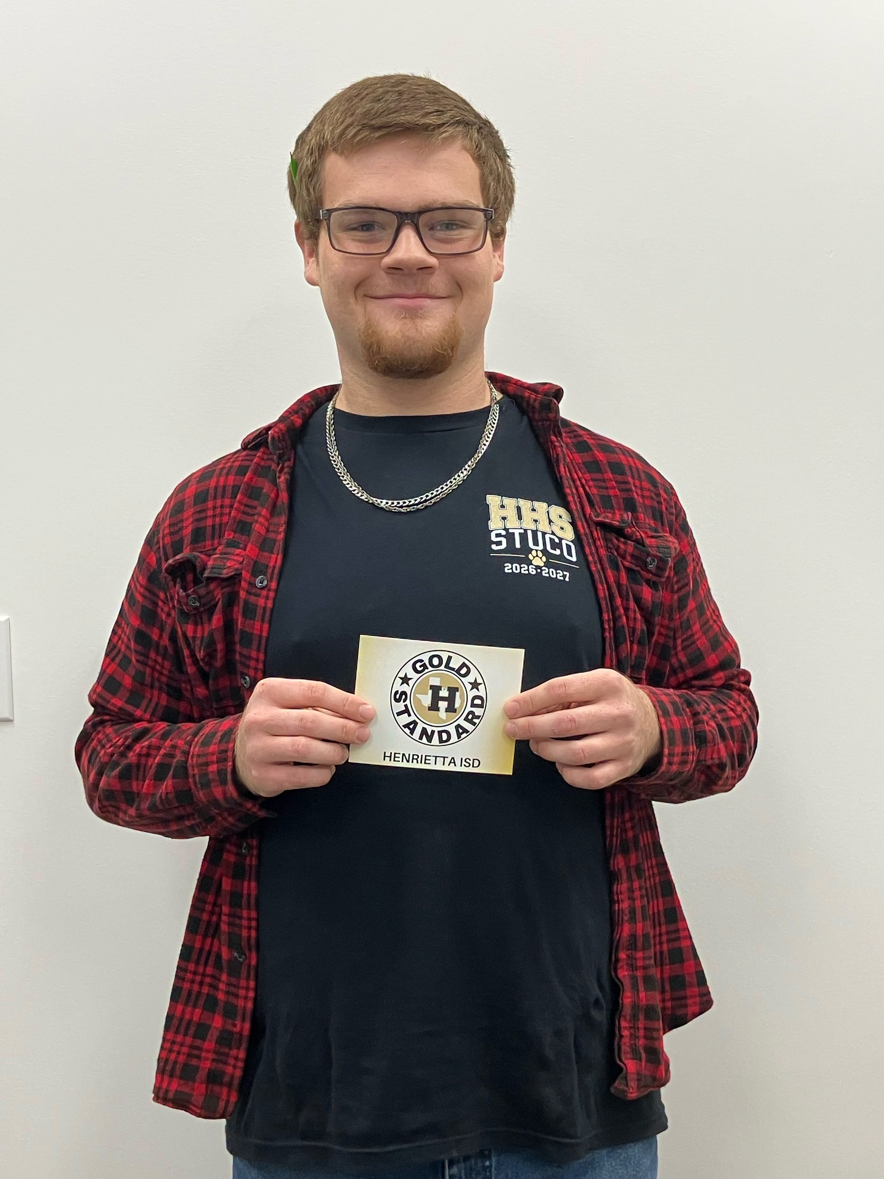 Man in glasses and necklace holds a small card, wearing a black shirt and red plaid jacket.
