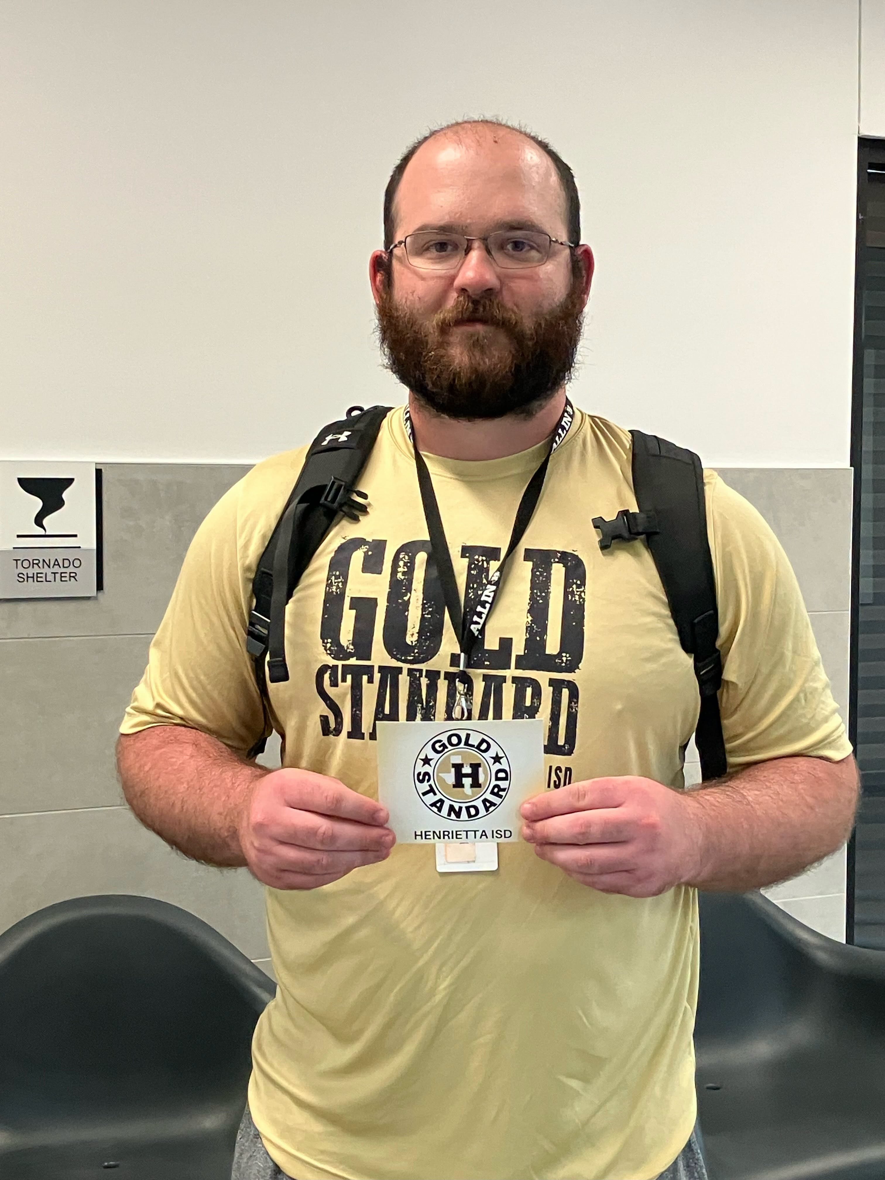A man in a yellow shirt holds a gold standard badge, wearing a backpack, with chairs and a white wall in the background.