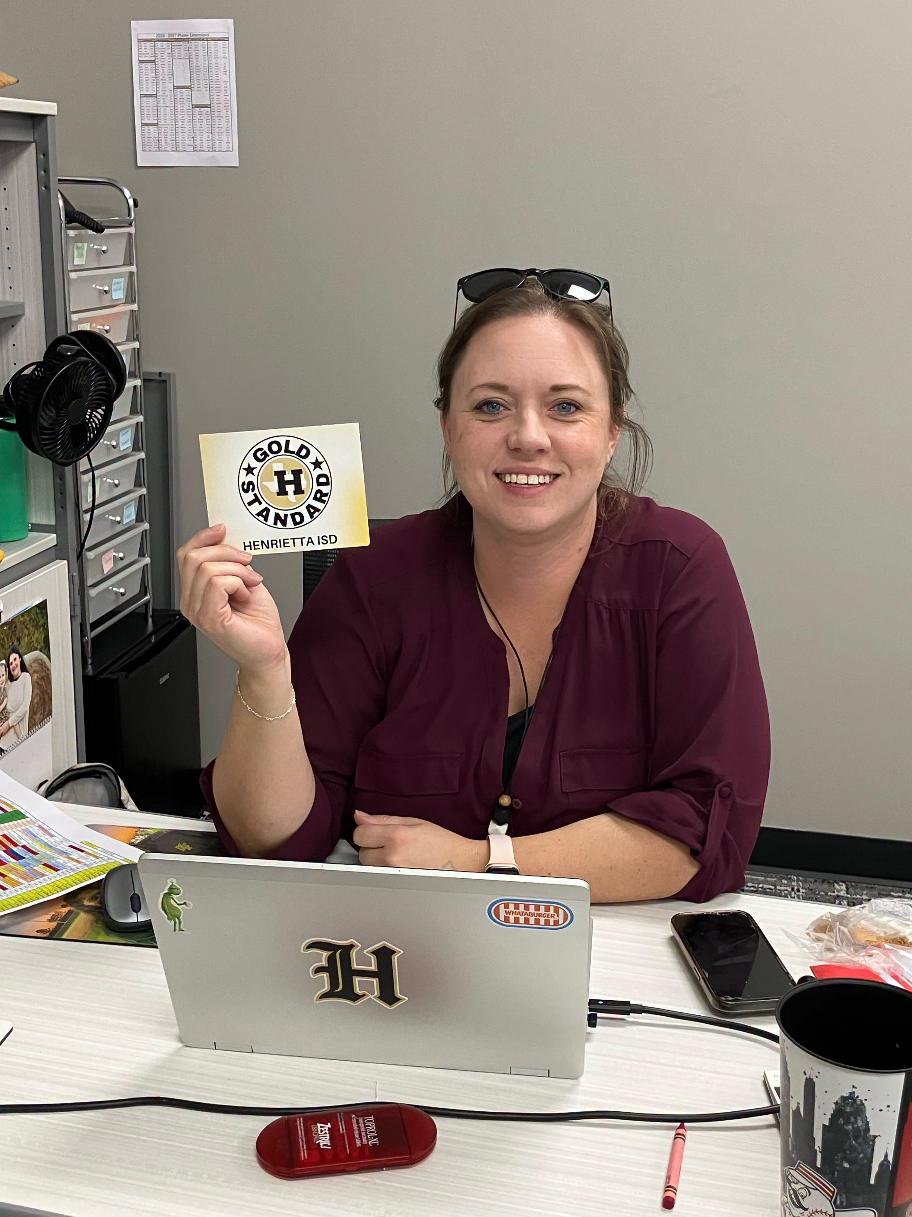 Woman smiling while holding a card at a desk with a laptop, cup, phone, and other items.
