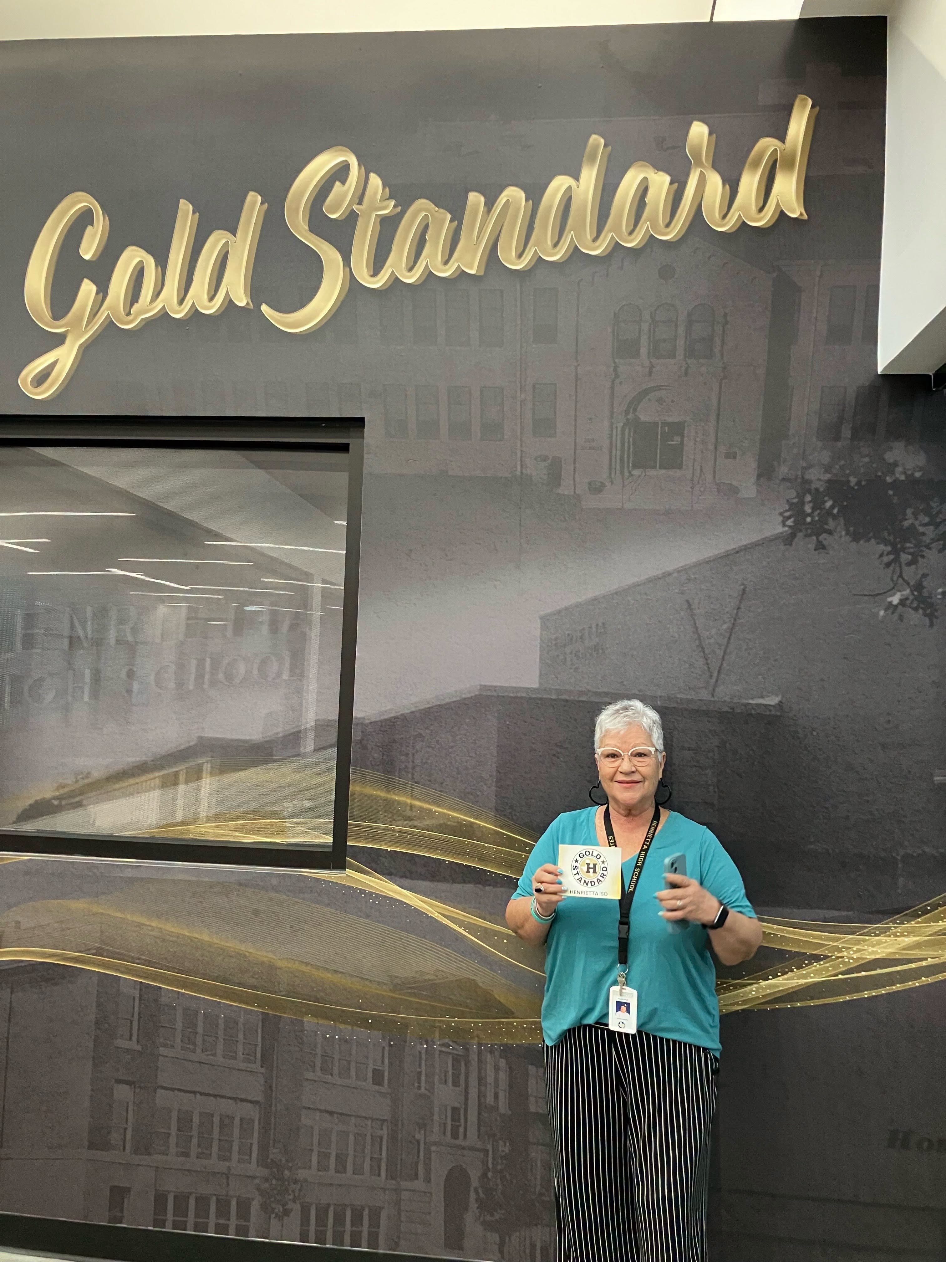 Person stands in front of a wall with "Gold Standard" text and buildings. They hold a card and wear a blue shirt and watch.