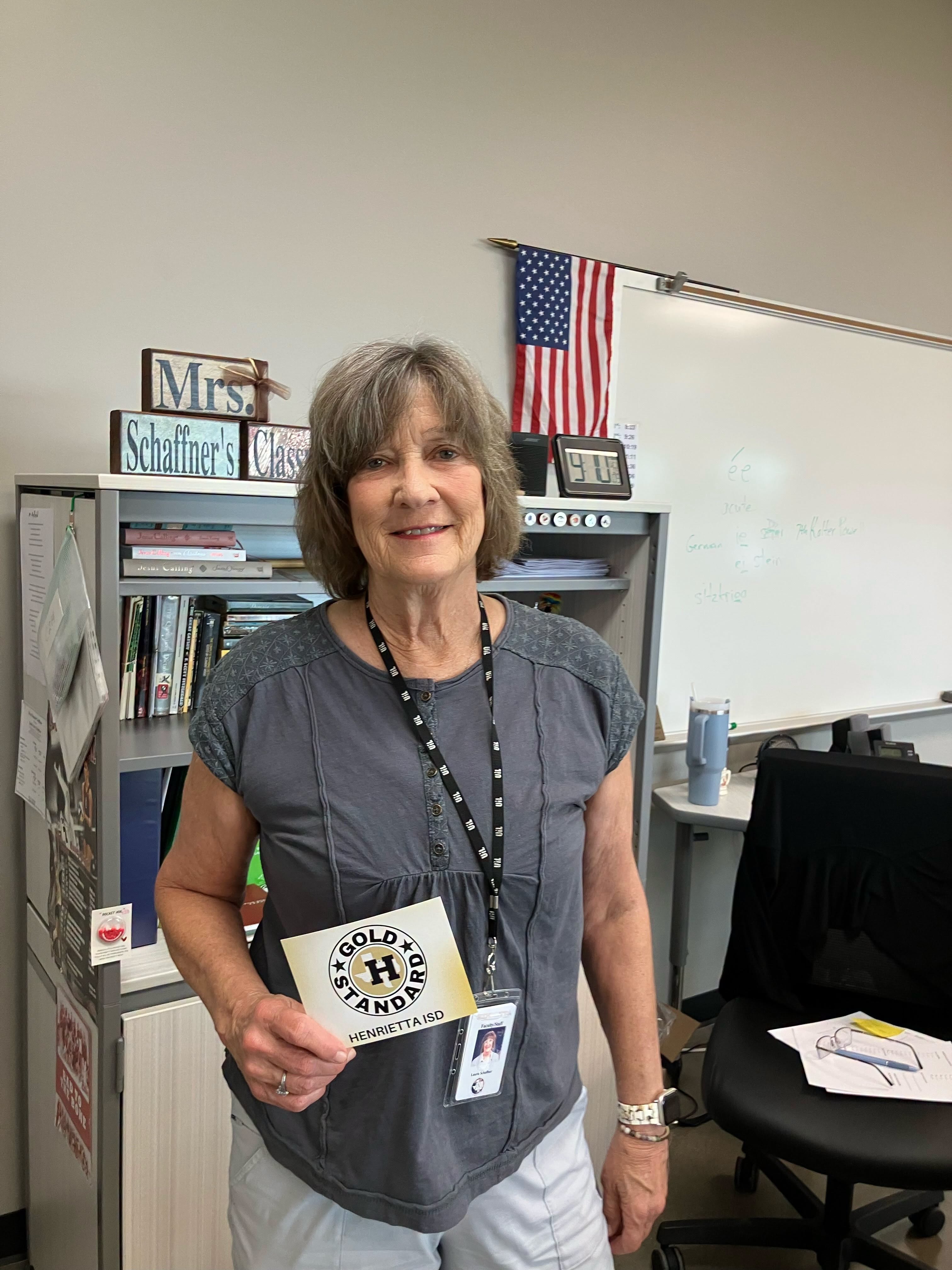 A woman in a classroom holds a card with "Gold Hands" text. She wears a name tag and smiles. Behind her is a bookshelf and a US flag.