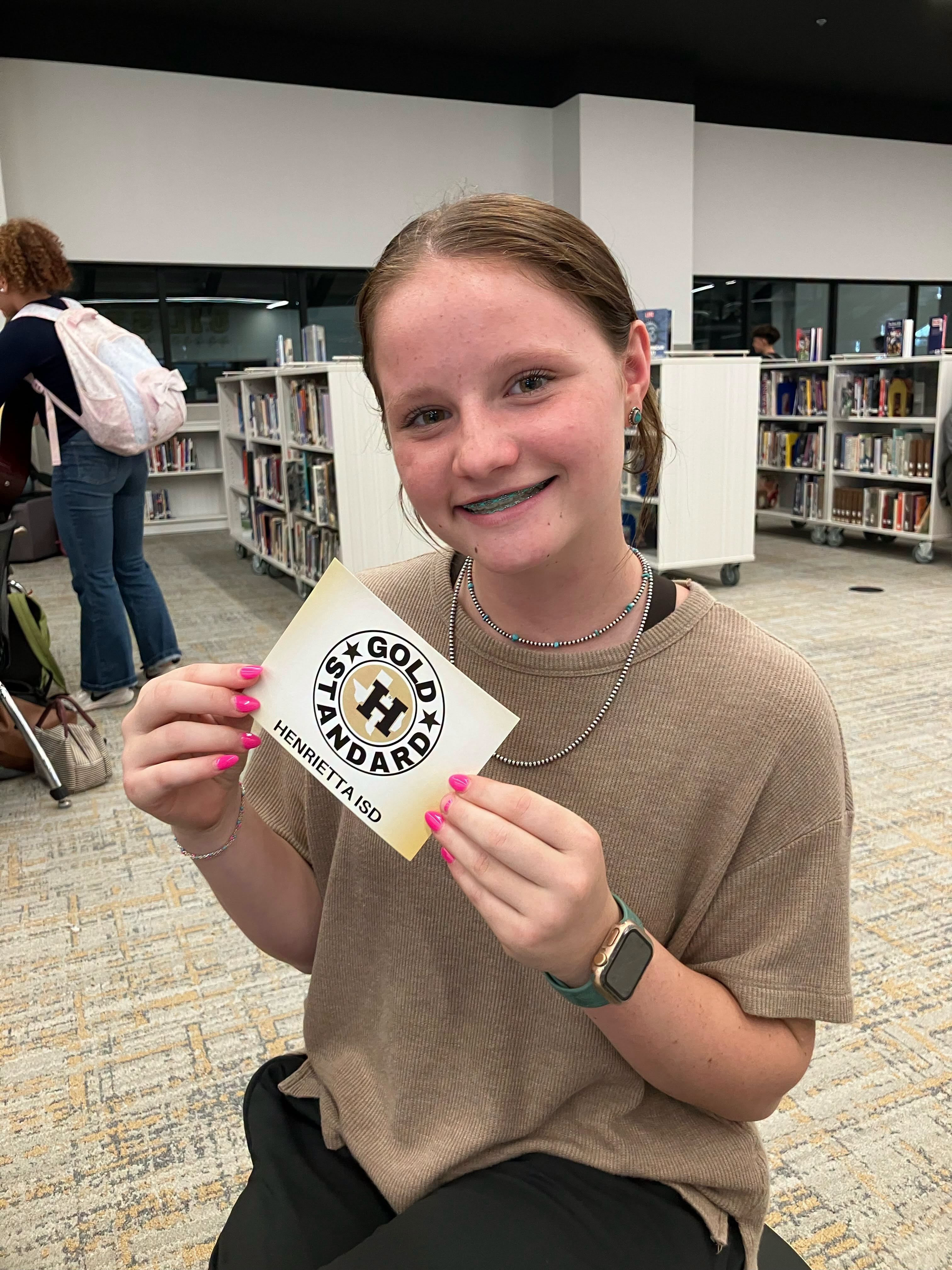 A girl holds up a yellow card with the words "HS GOLD HANDBOOK" in a library.