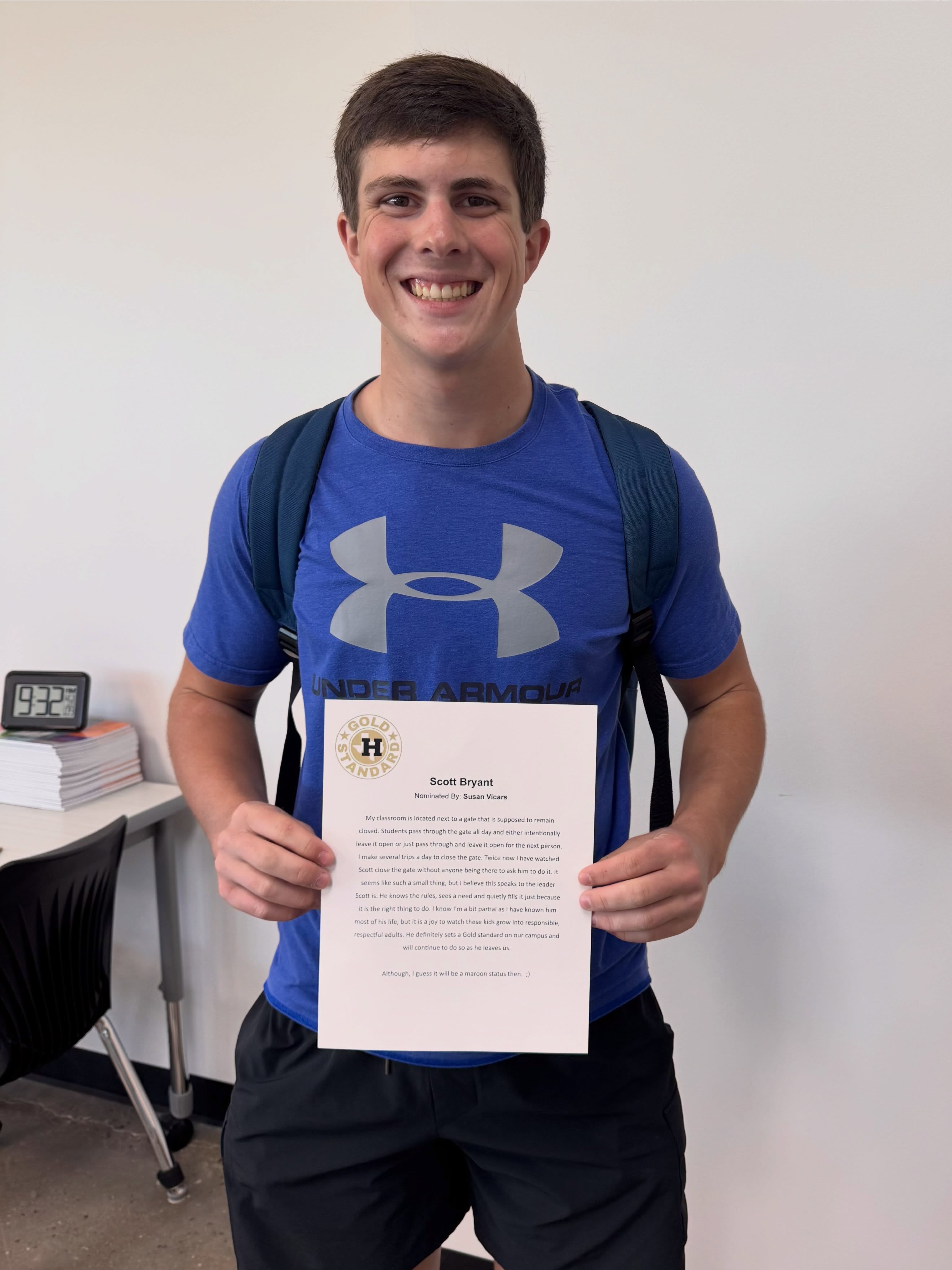 A smiling man in a blue Under Armour shirt and black pants holds a certificate. Behind him is a desk with books and a clock.