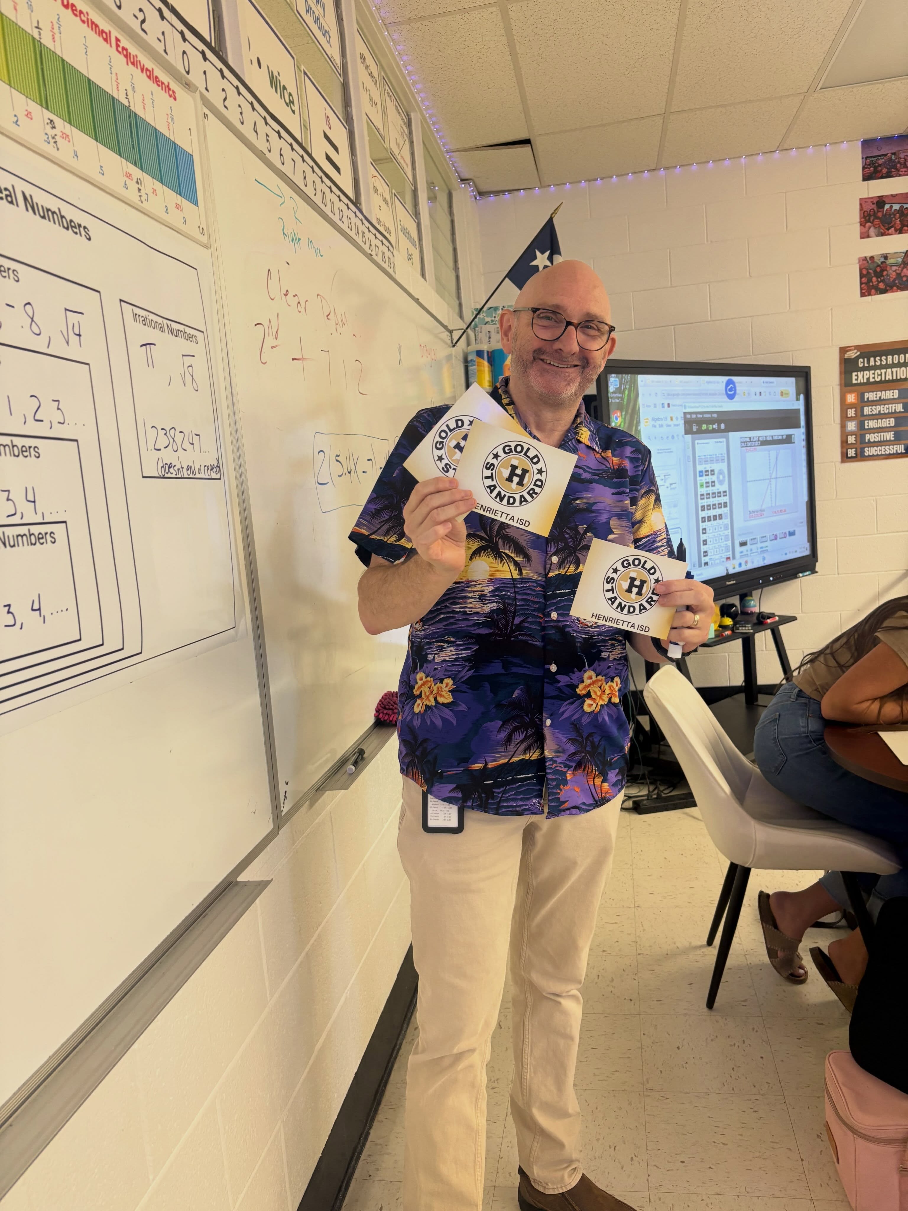A man in a patterned shirt holds cards in front of a whiteboard with numbers. A monitor, flag, and chairs are in the background.