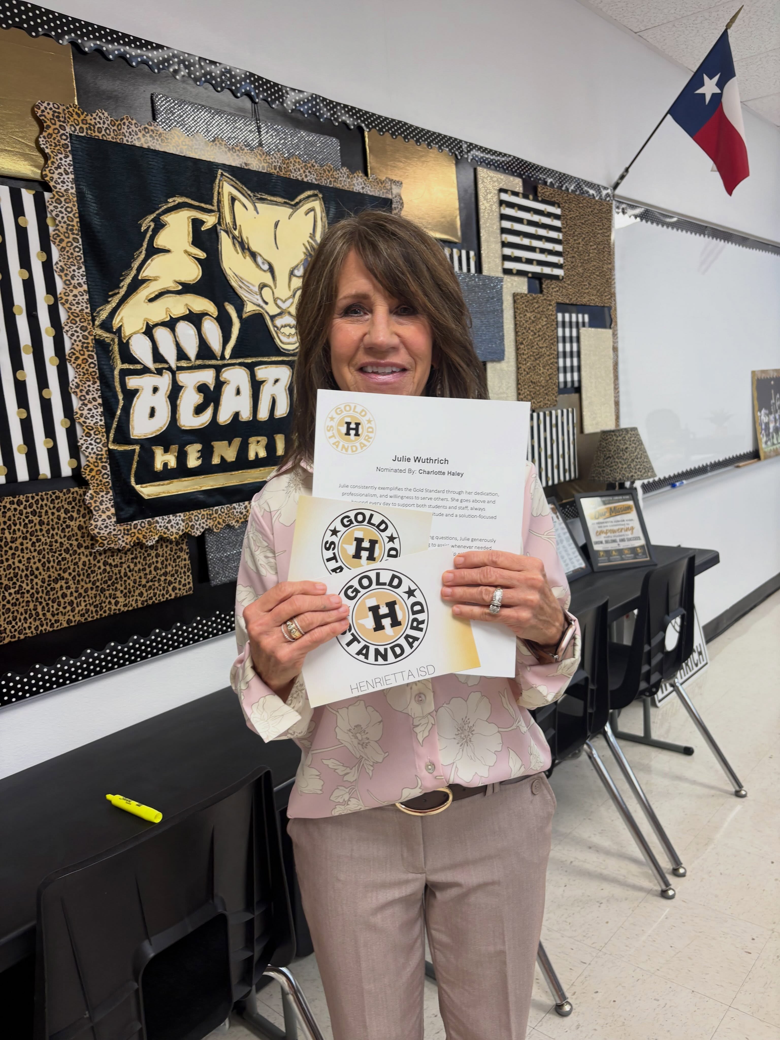 A smiling woman holds a certificate in a classroom with a wall backdrop, a Texas flag, and black tables.