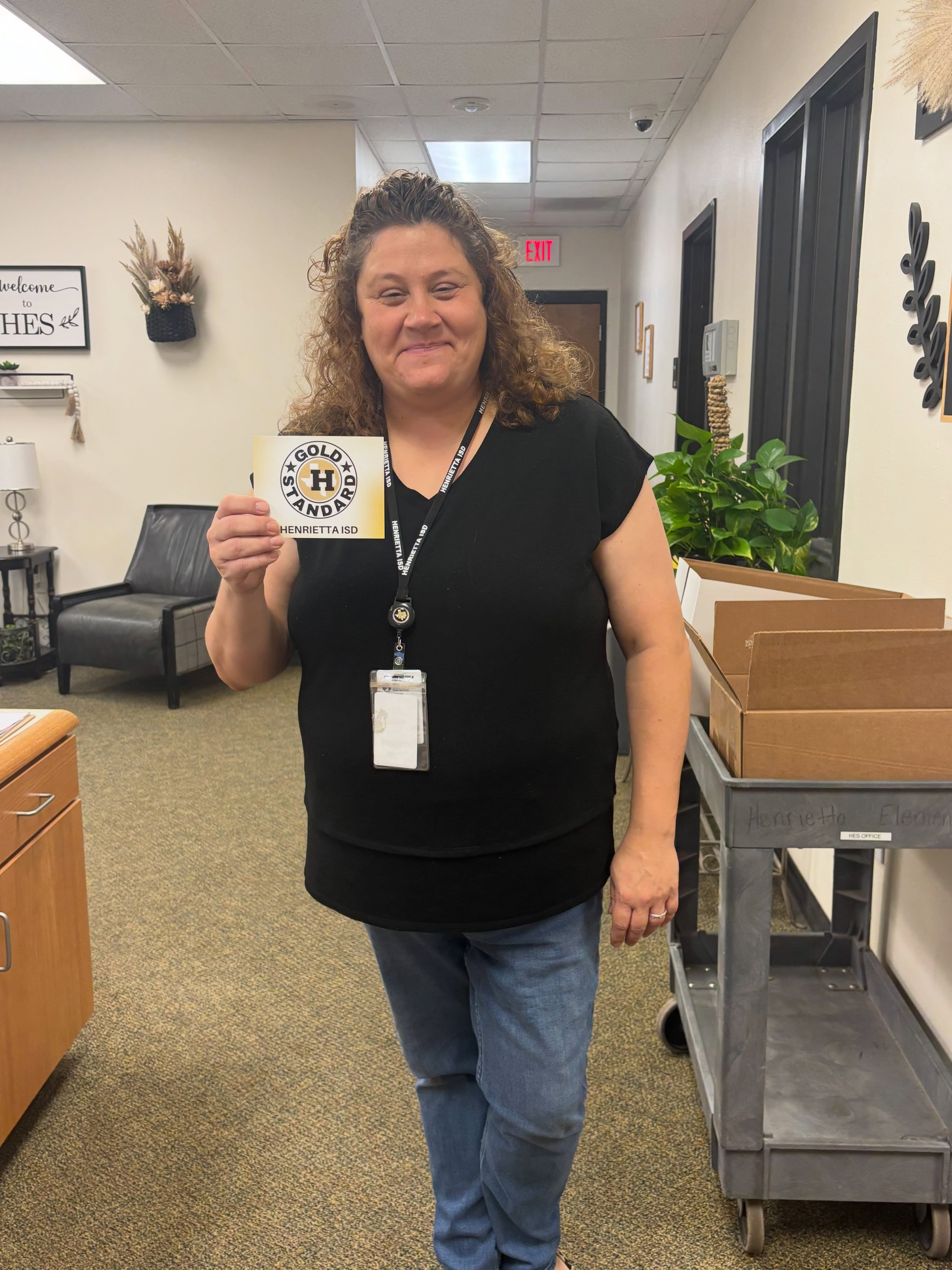Woman in an office hallway holds a card and smiles. Behind her, there is a metal cart with boxes.