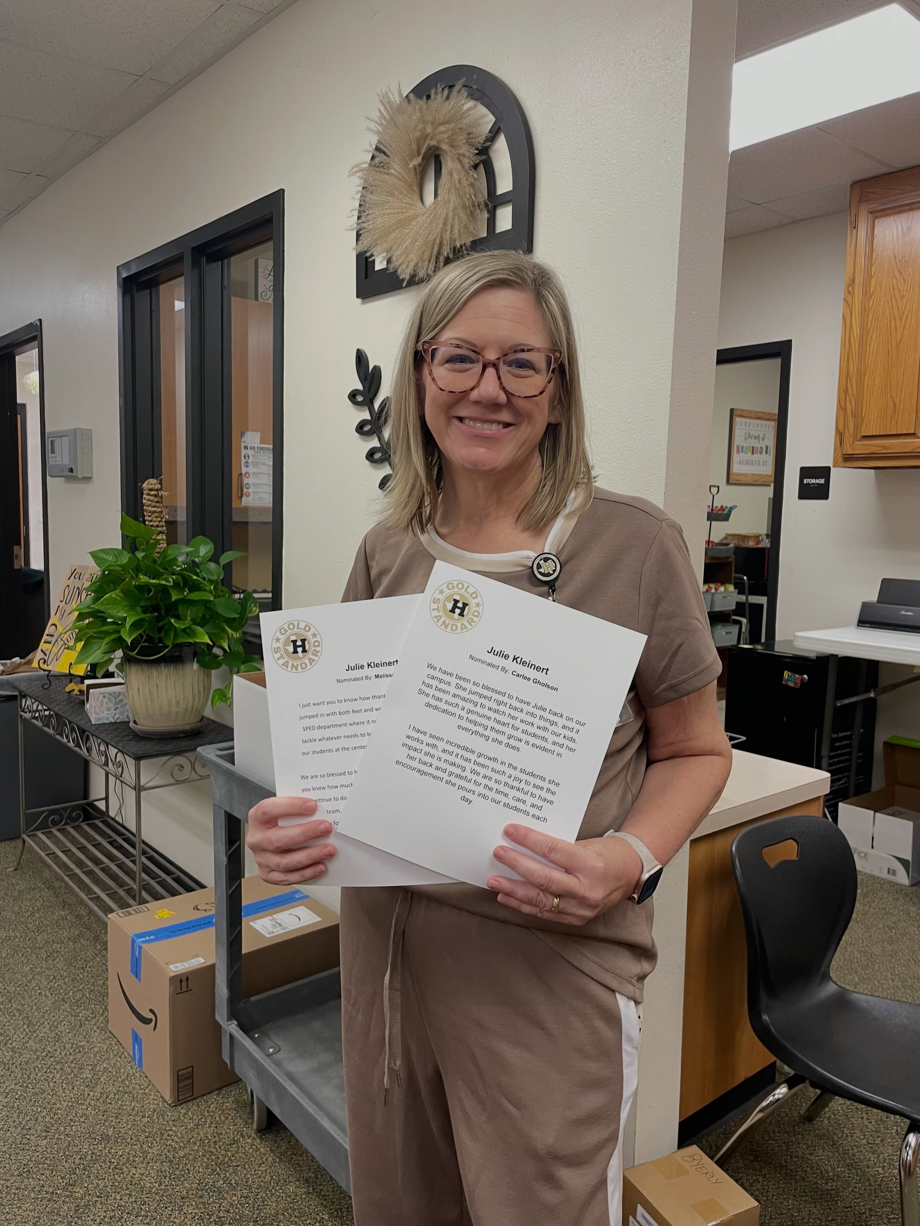 A woman wearing glasses and a brown shirt holds a certificate with black text in both hands.