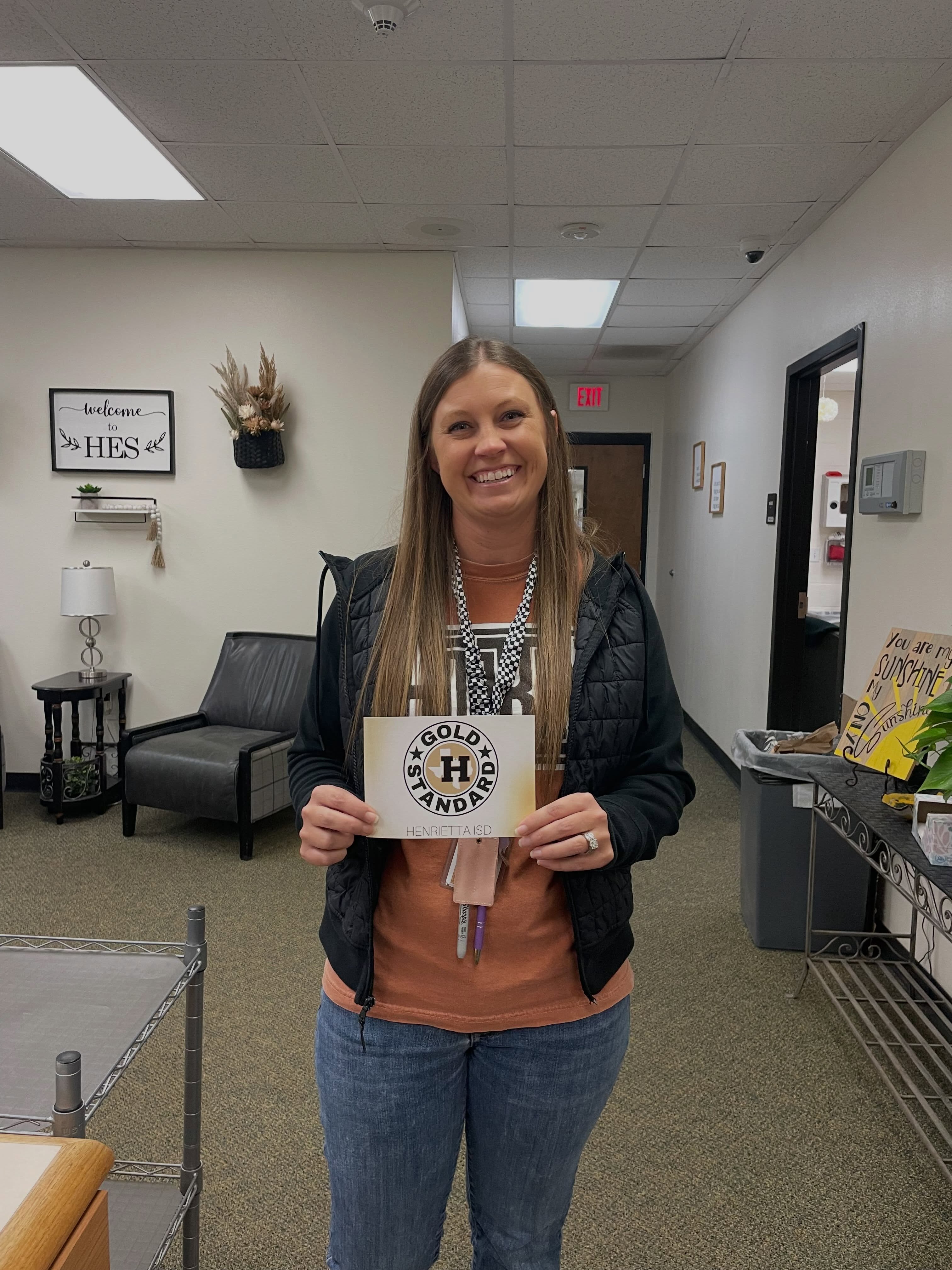 A woman stands in a hallway, holding a sign that reads "GOLD HAND". She wears a jacket and jeans.