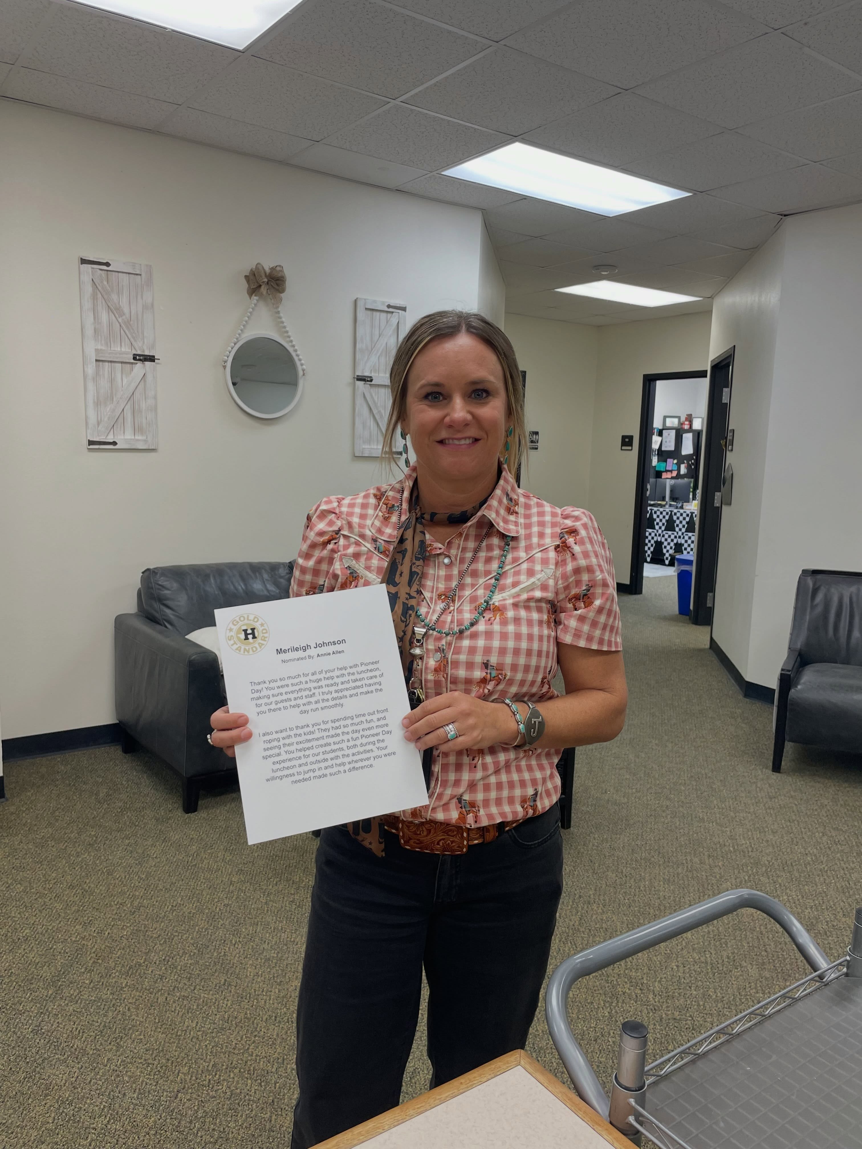 Woman in checkered shirt holding certificate in hands. Background includes hallway, couch, mirror, and ceiling lights.