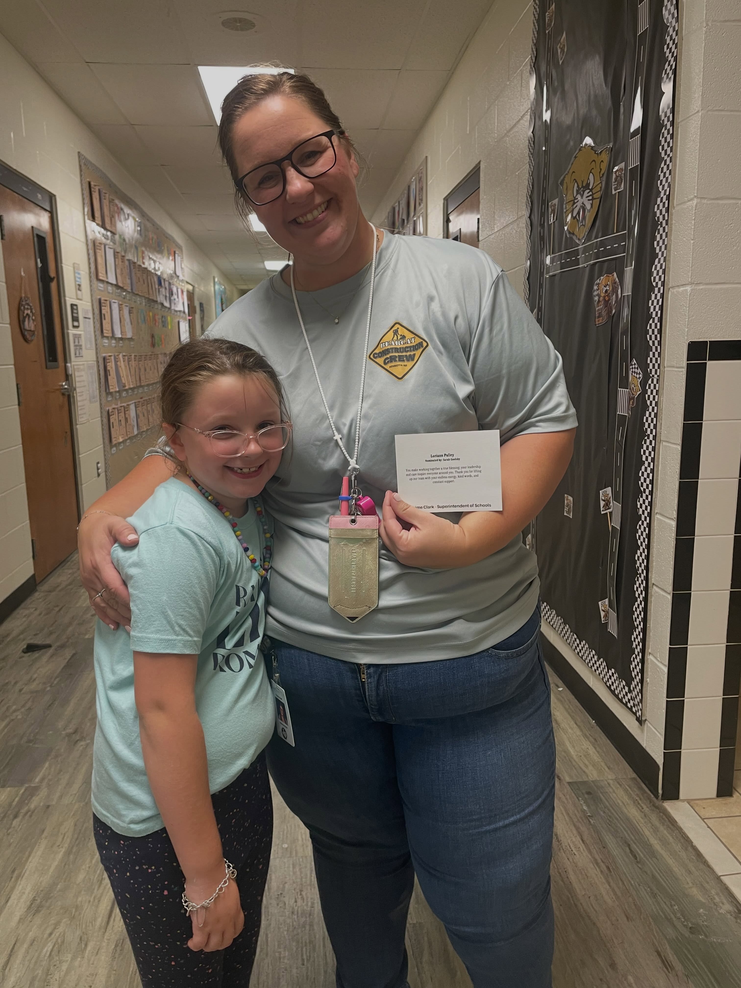 Two people standing in a hallway. A woman holds a certificate and a child smiles. They are near a wall with black and white artwork.