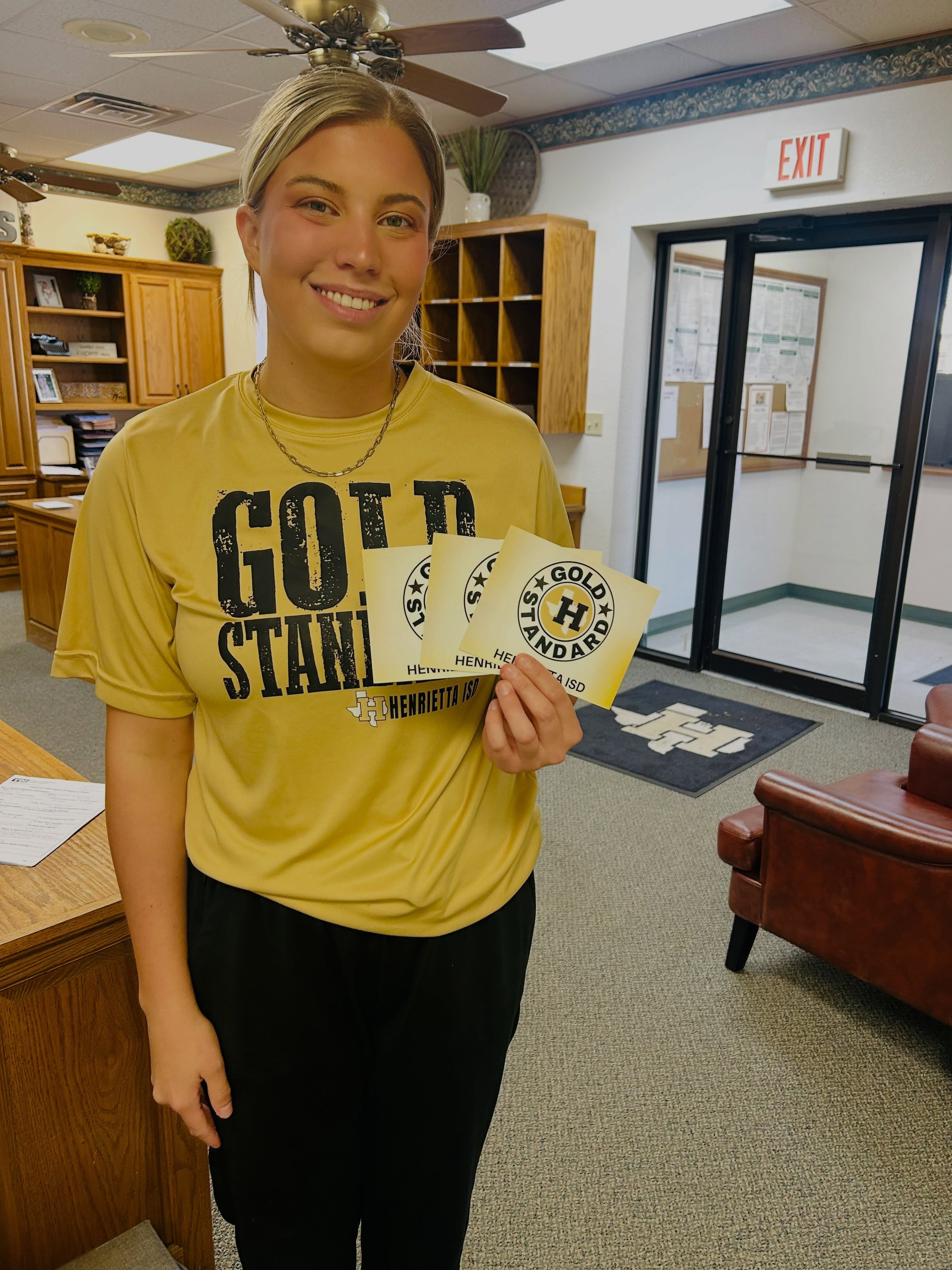 A woman in a yellow shirt stands holding cards, smiling. Behind her is a desk, bookshelves, and an exit sign.