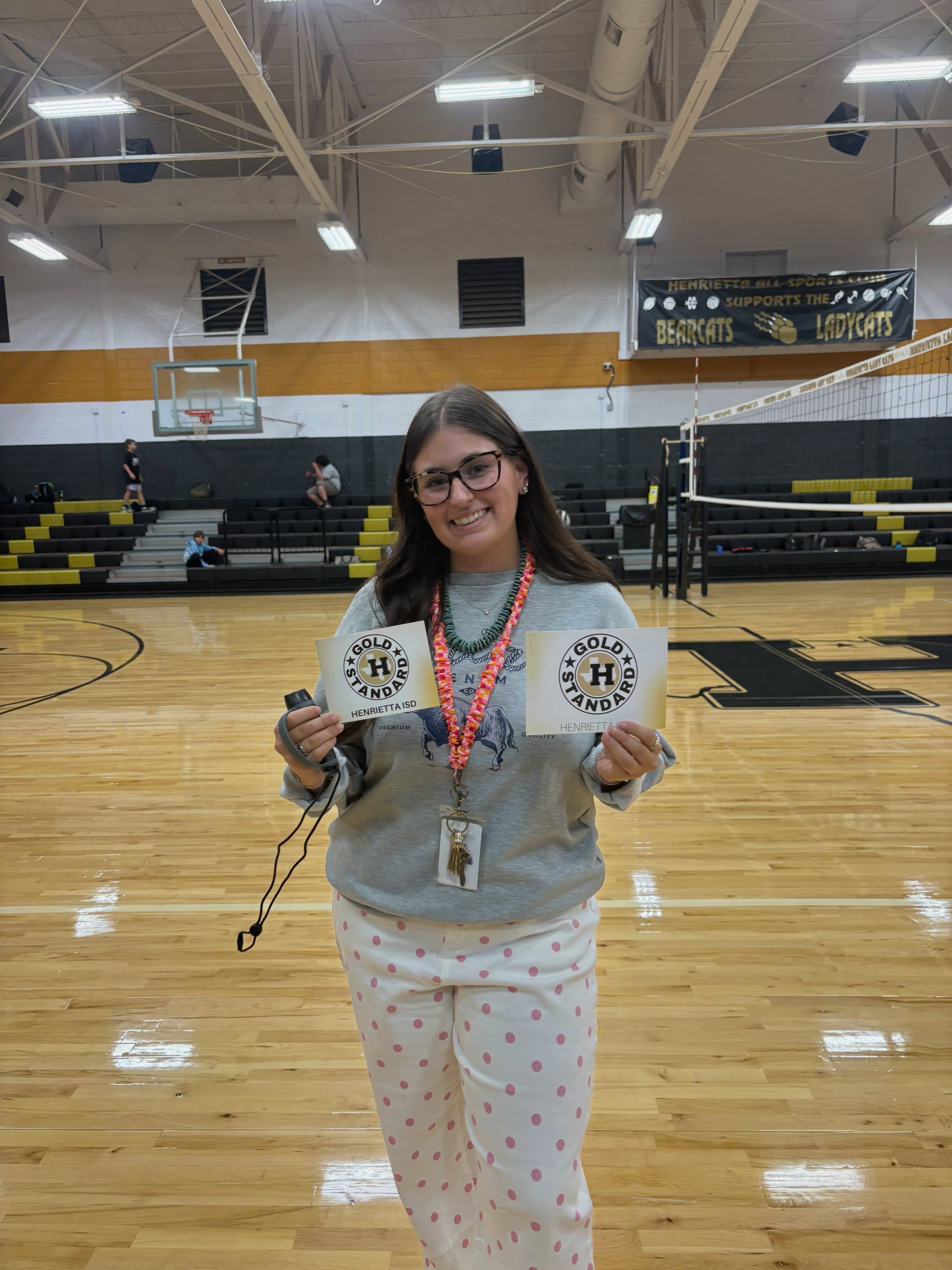 Person wearing glasses and a sweatshirt stands in a gymnasium holding two signs with a logo.