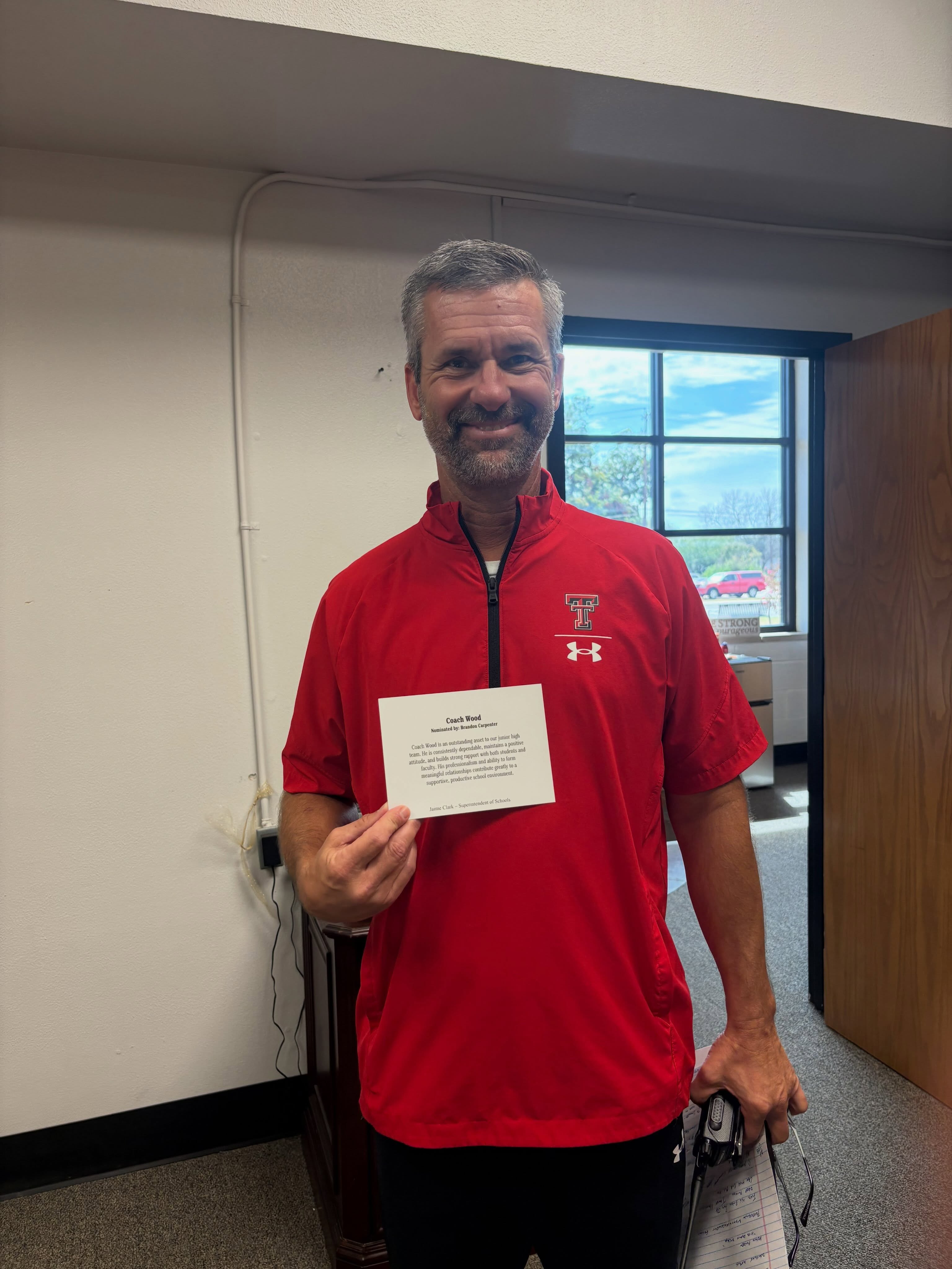 A man with a smile, holding a white card, wearing a red shirt with an Under Armour logo.