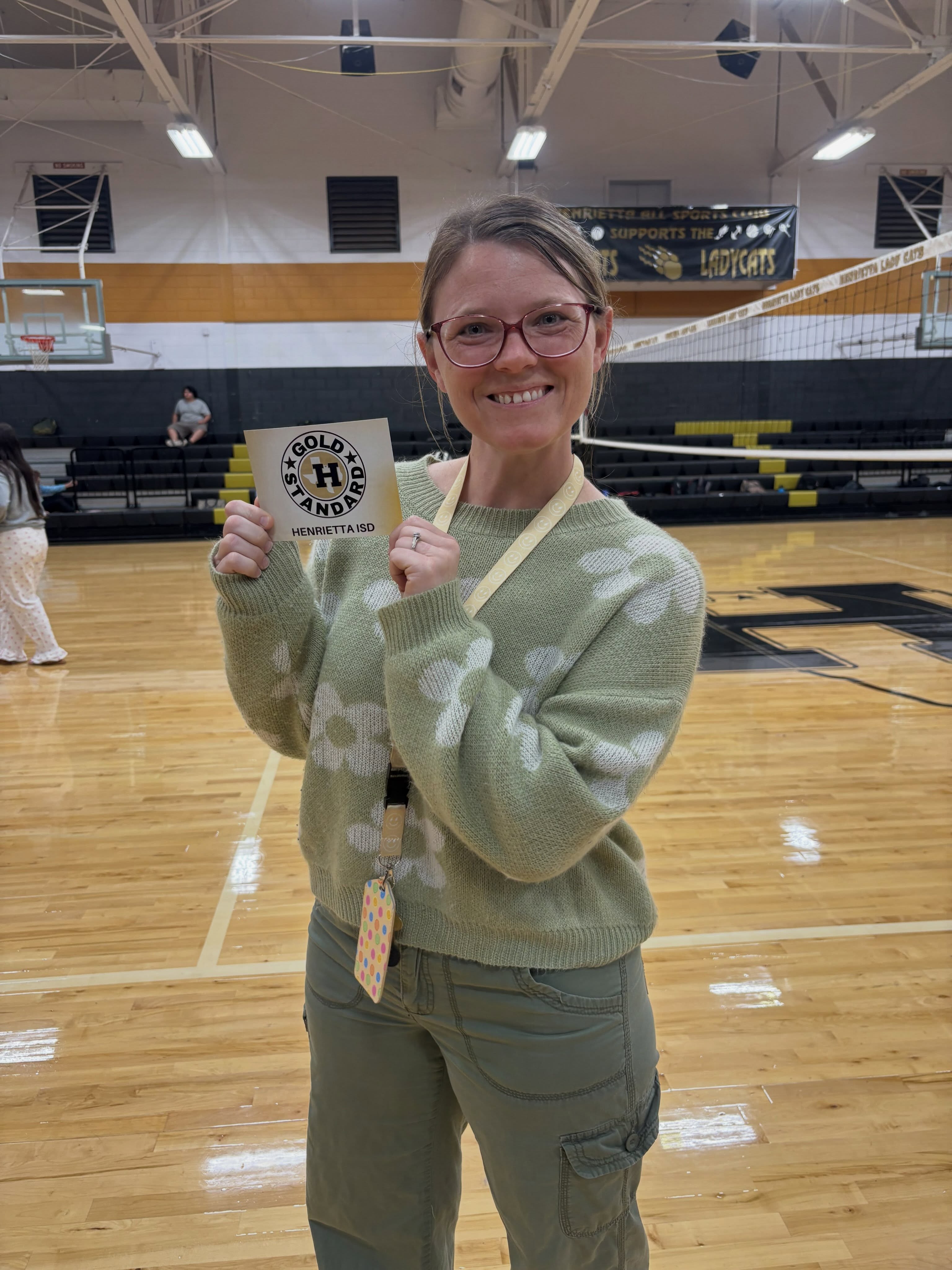 A woman in glasses and a camouflage sweater holds a card and ID on a basketball court.