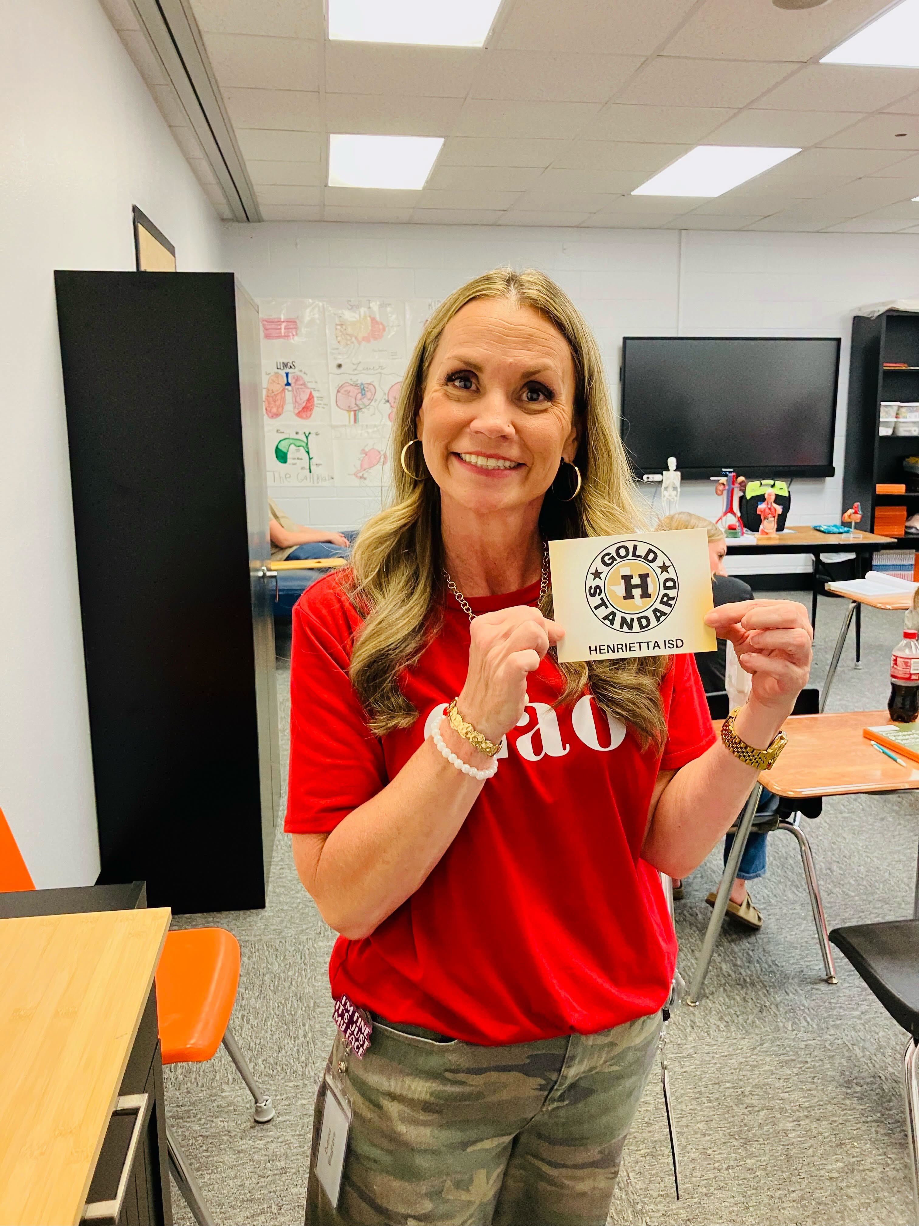 Woman in red shirt holding up a card with "Houston Gold Hands" written on it in a classroom.