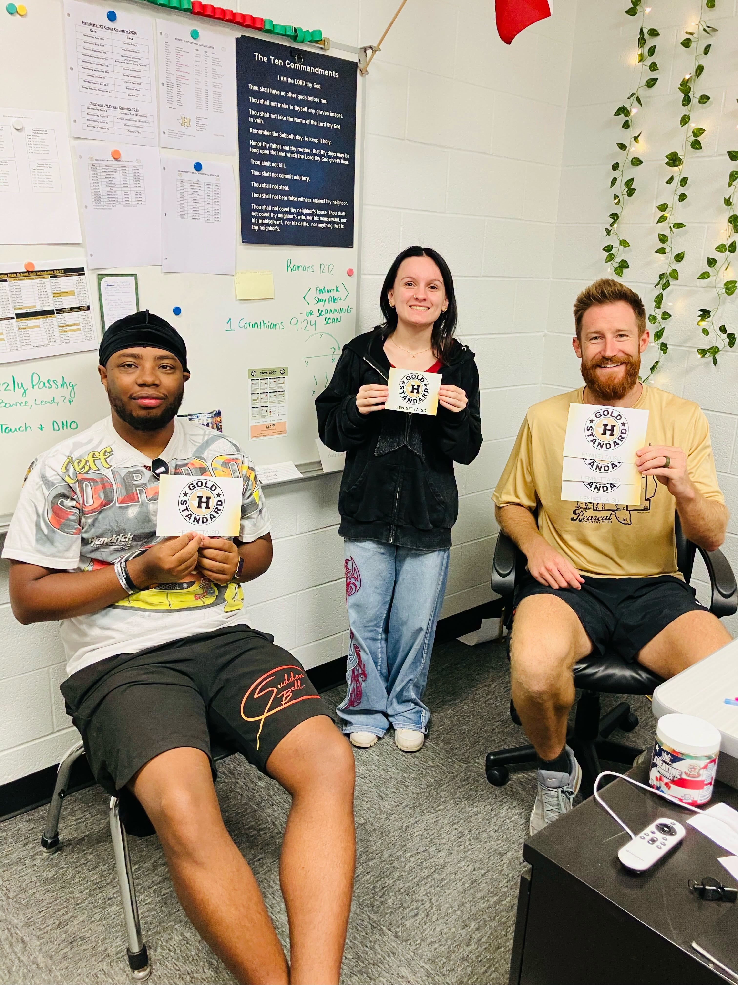 Three people hold certificates and smile, seated in an office. A whiteboard and papers are on a wall behind them.