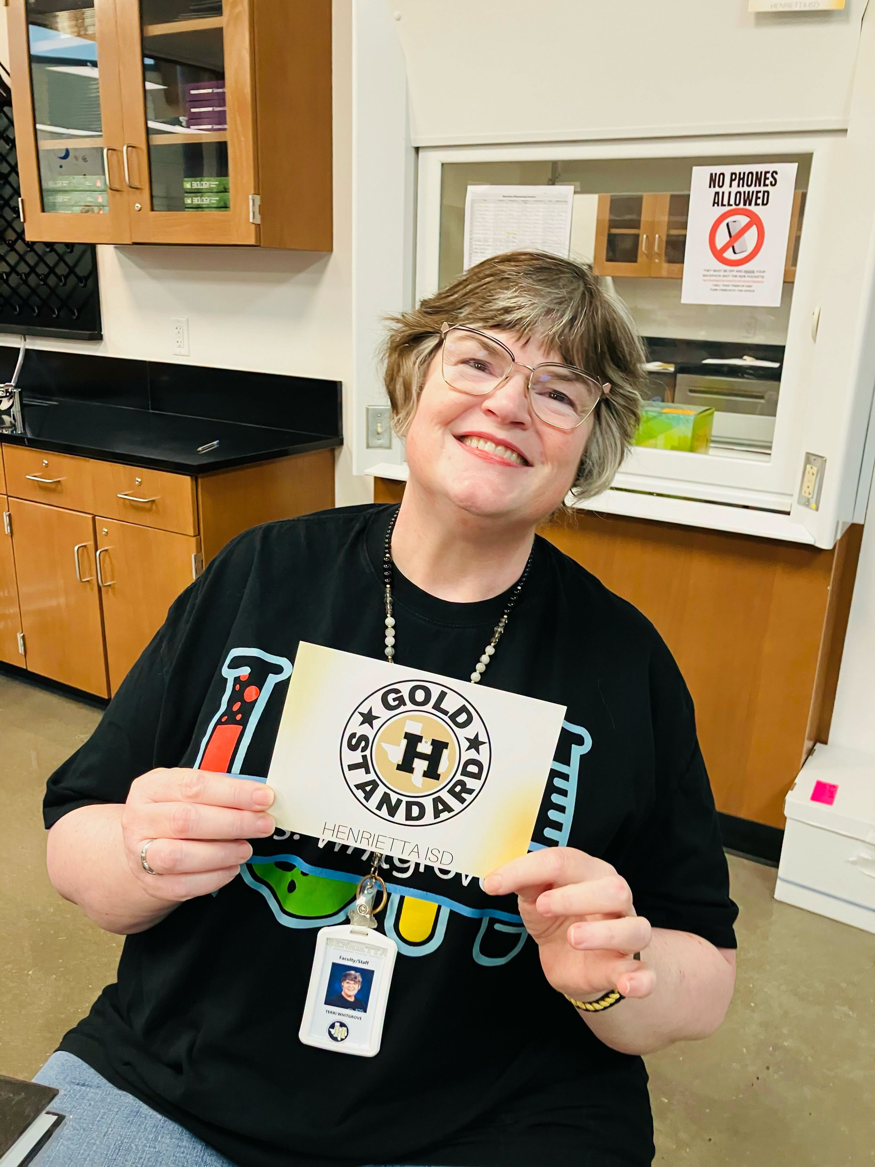 Person in black shirt sits in a lab. They hold a gold standard badge, smiling. Lab equipment and cabinets in the background.