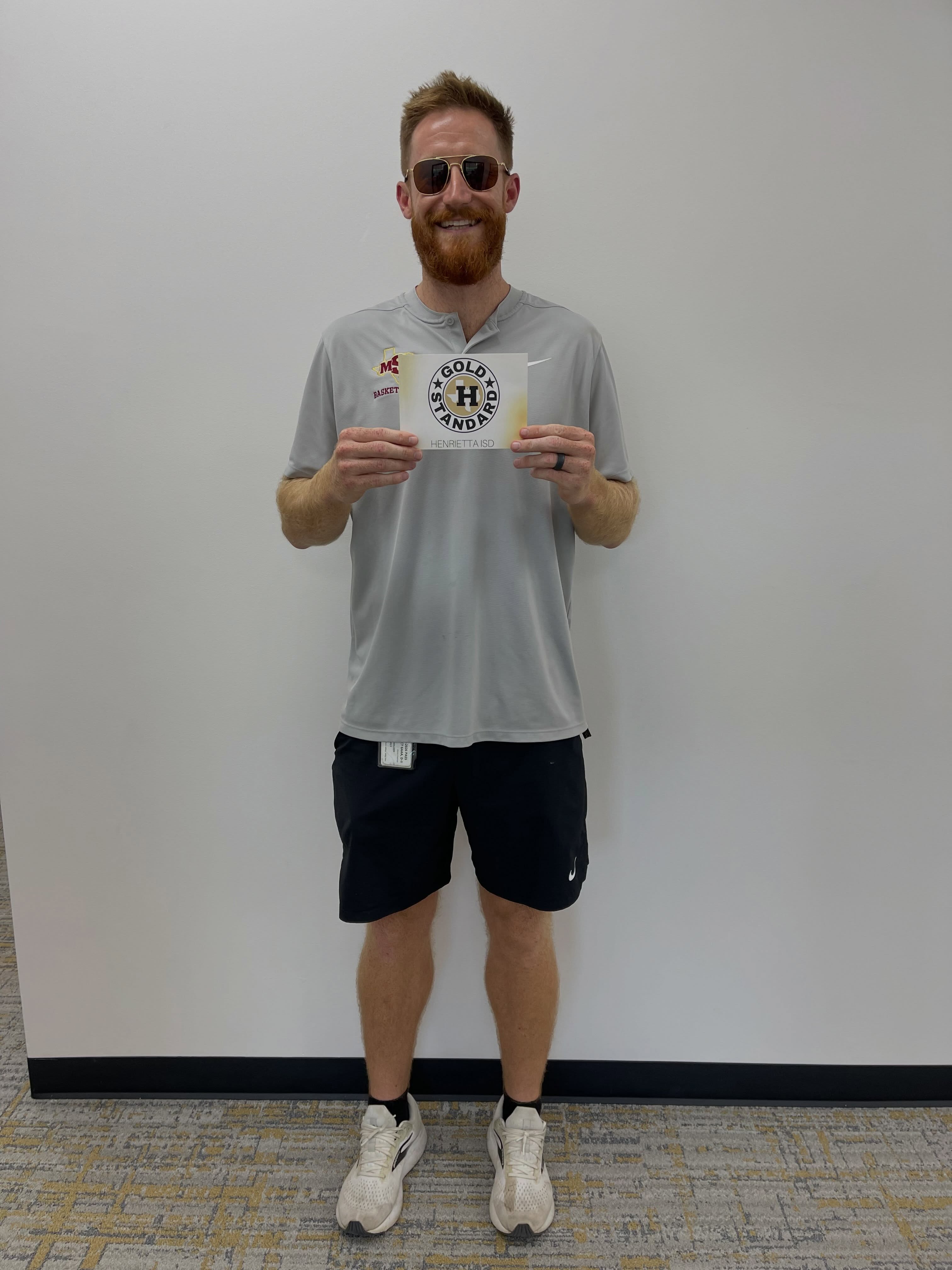 Man in a gray shirt and black shorts holding a card with text and logos stands against a white wall.