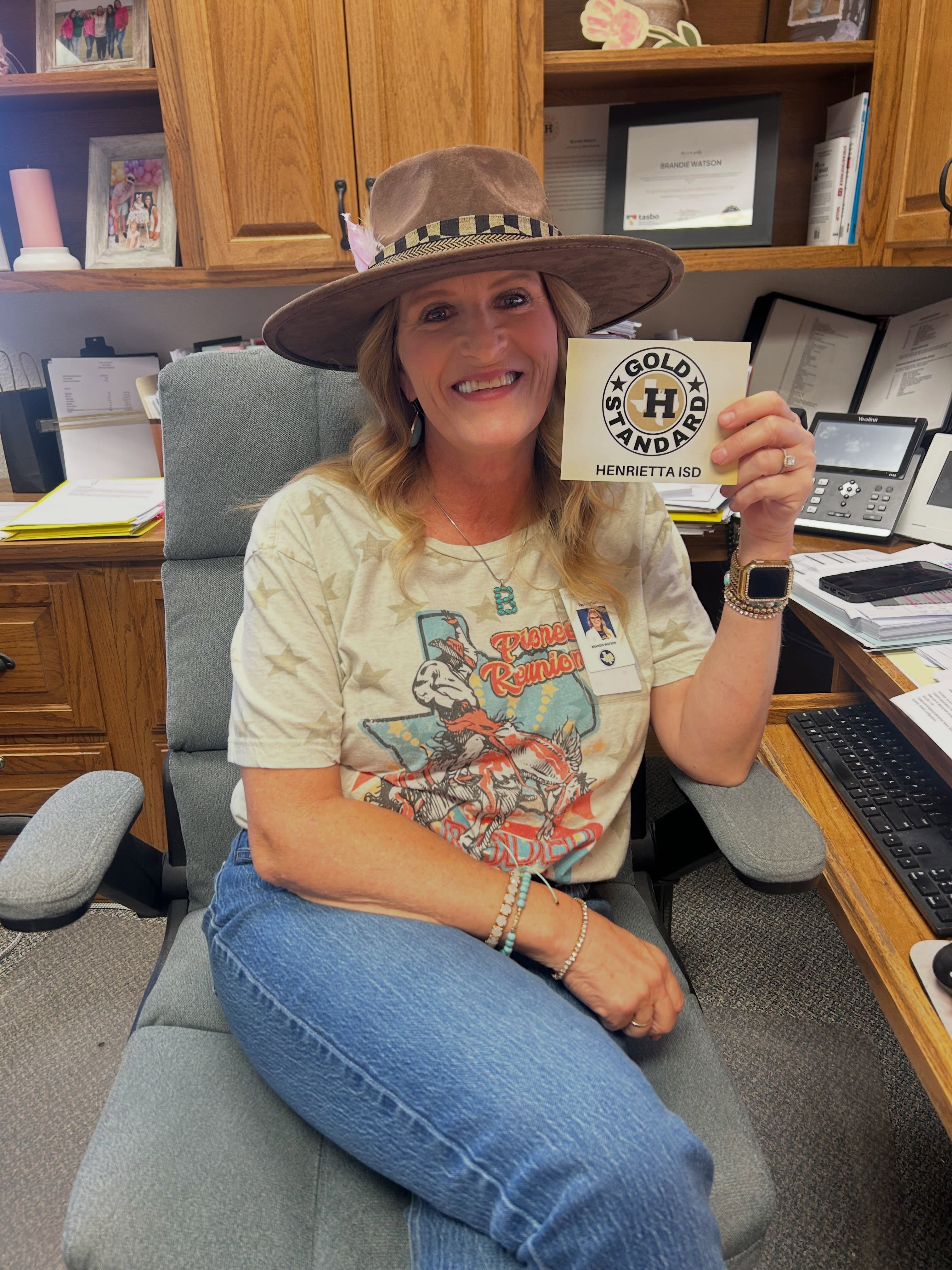 Woman in a hat holds a small card with a smiling face. Behind her, an office environment with wooden cabinets and a computer.