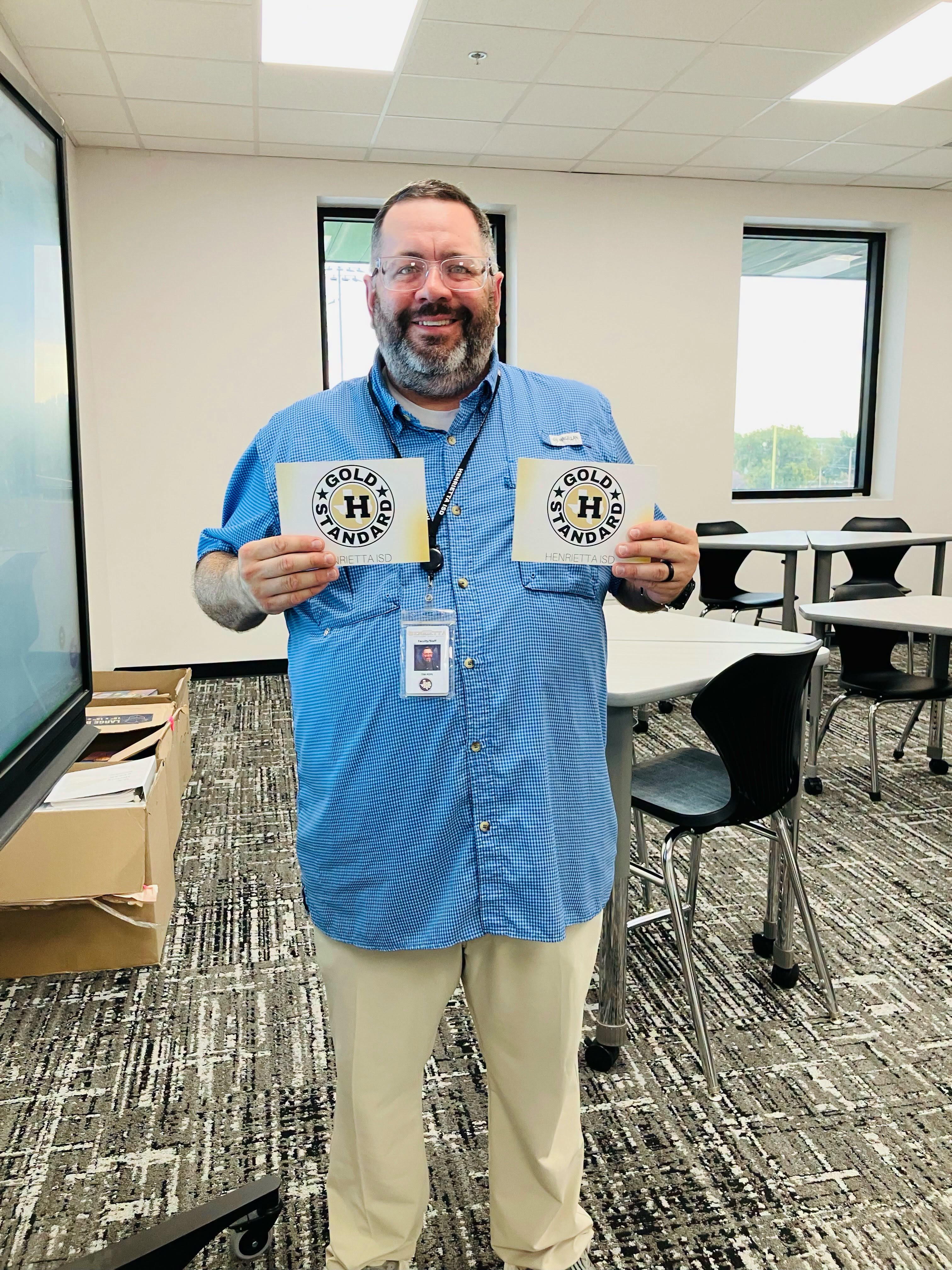 Man in a blue shirt holds two white cards with the text "GOLD HAND". He is in a room with chairs, tables, and a monitor.