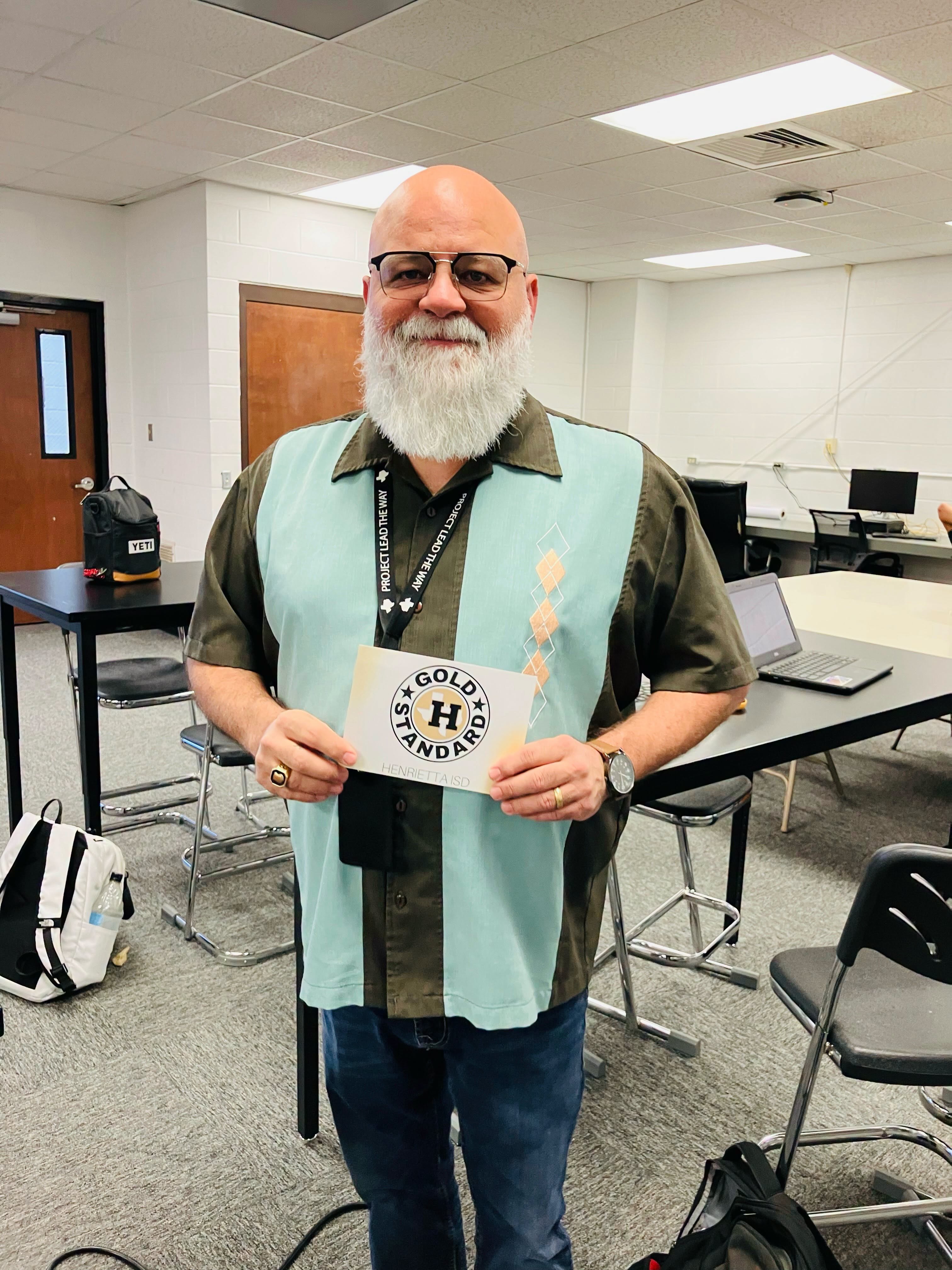 A man with a white beard and glasses holds a white certificate in a classroom. He wears a green shirt and a blue vest.