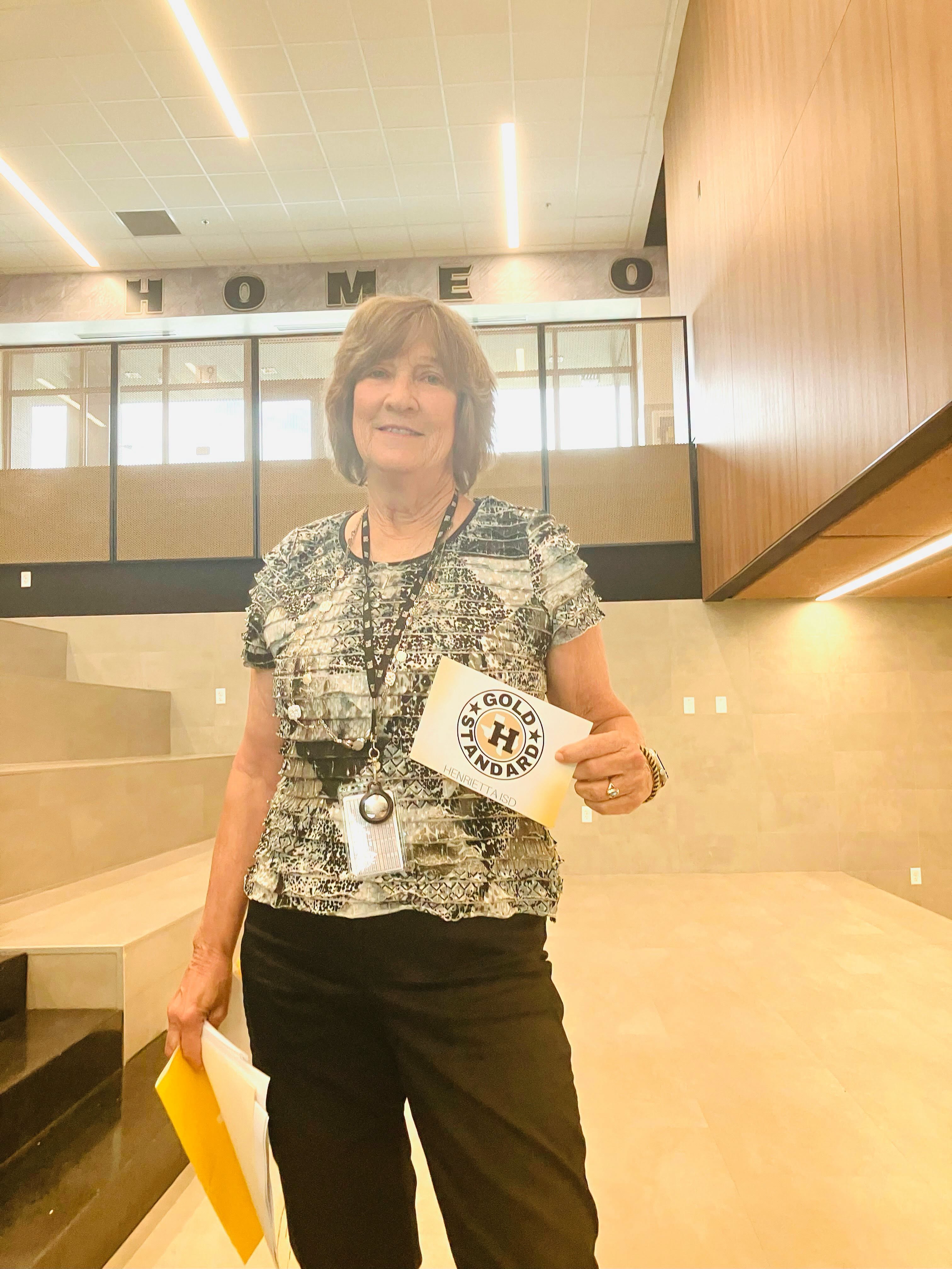 Person holds folder and sign with "GOLD BAND" text. Person stands in a room with a wooden wall and glass ceiling.