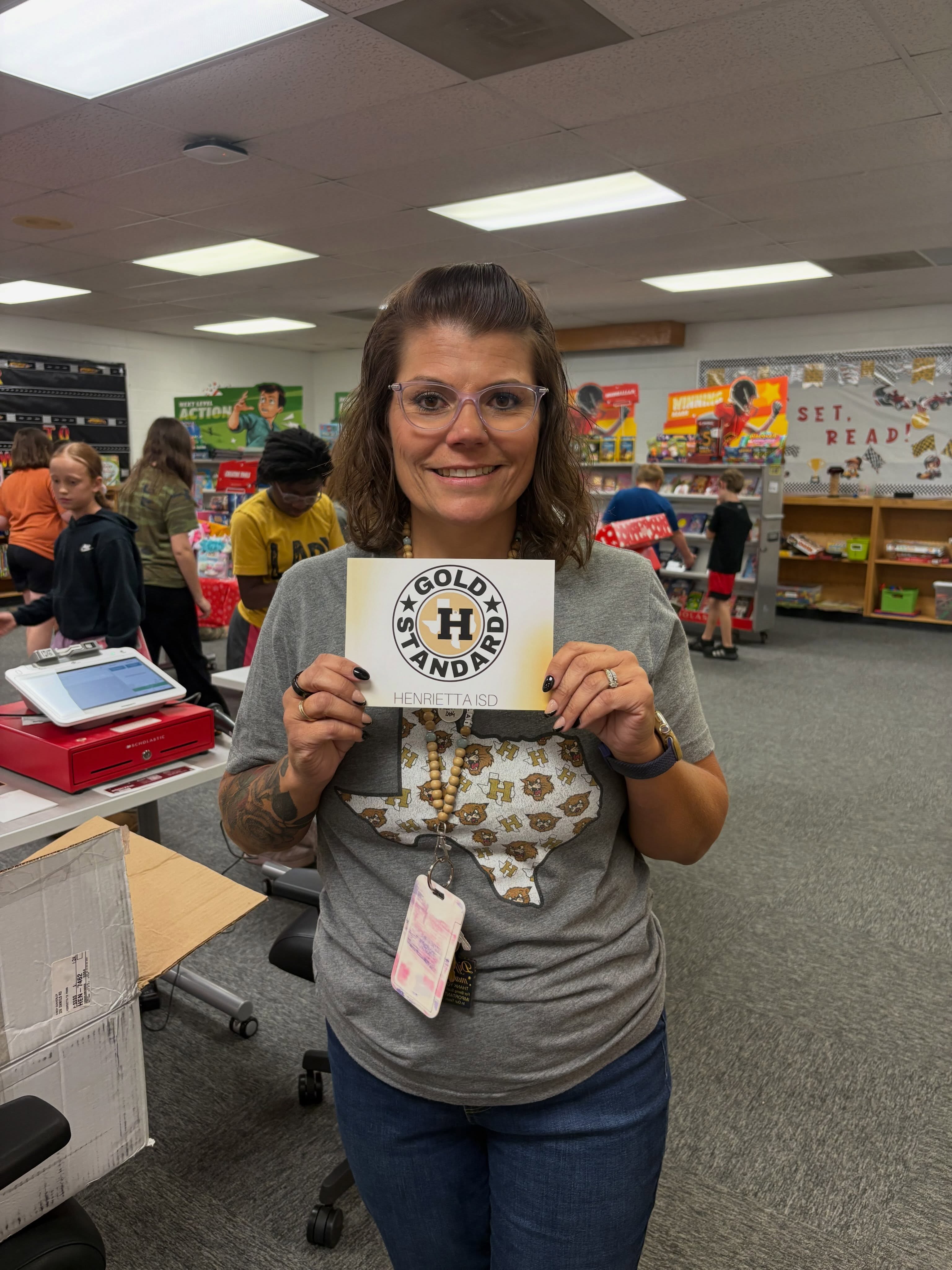 A woman smiles while holding a paper sign, standing in a classroom. She wears a gray shirt, glasses, and a necklace.