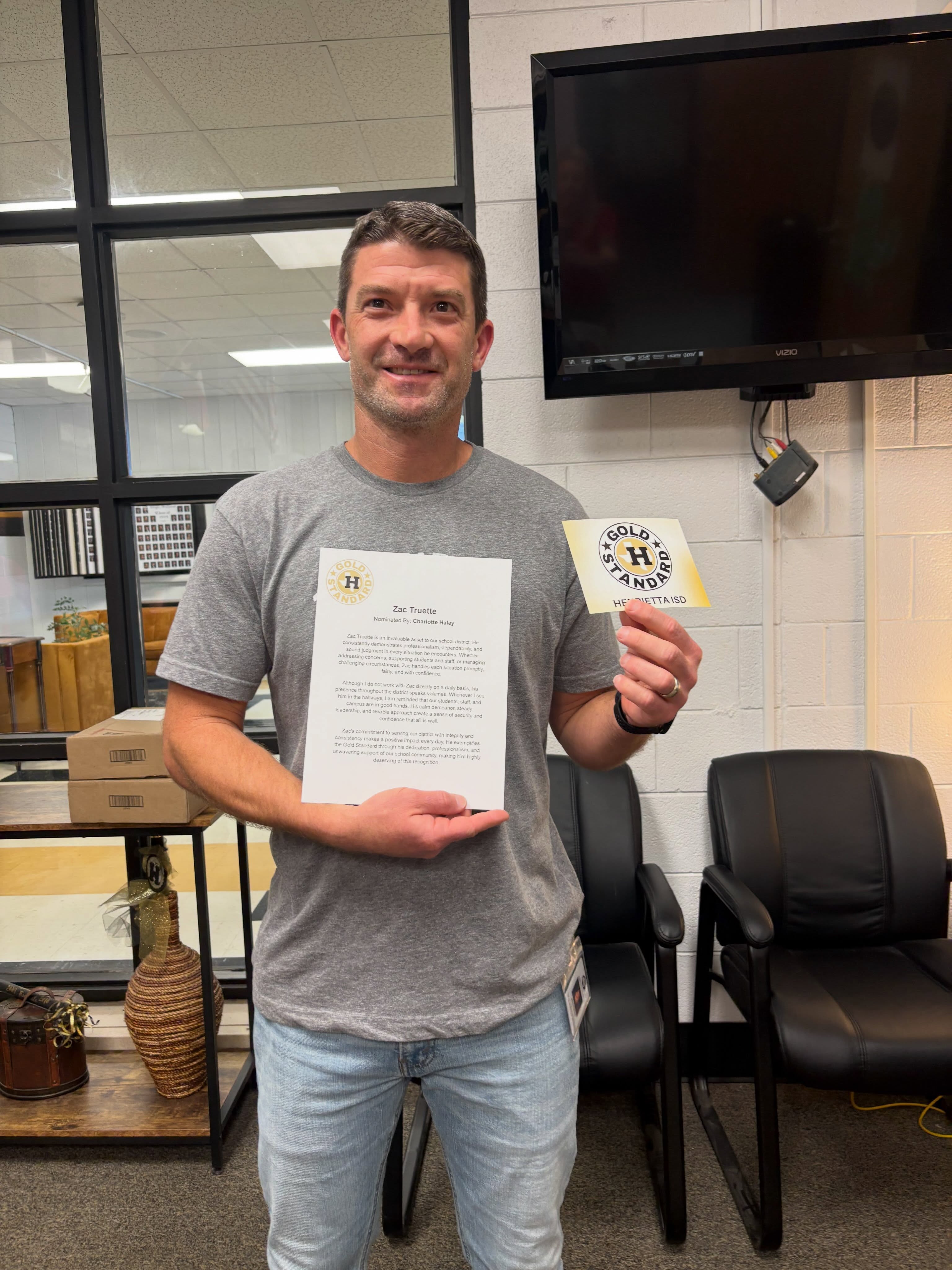 A man stands in a room holding a certificate and a card. Behind him, a wall has a TV and a mounted power outlet.