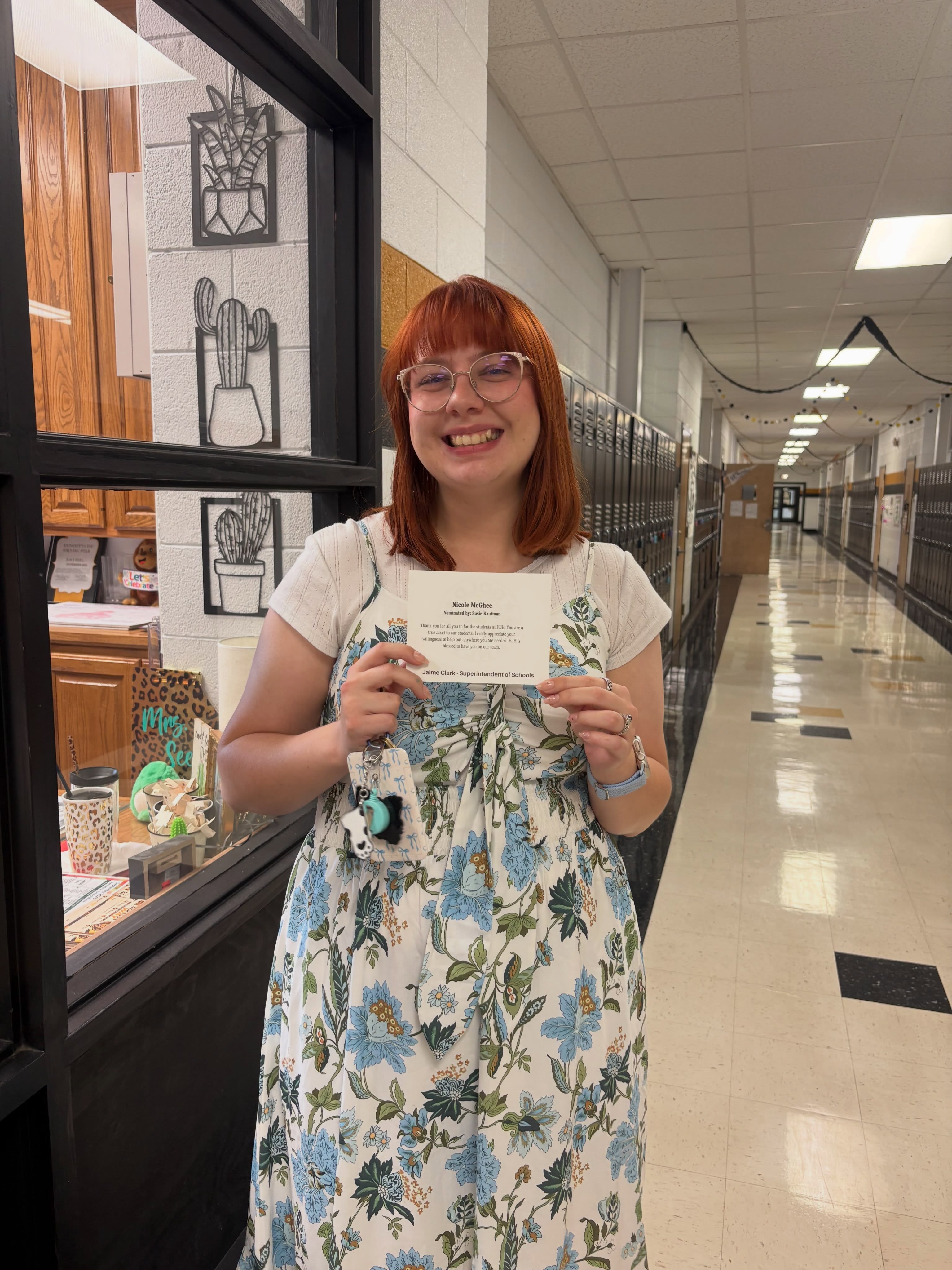 Person wearing glasses and a floral dress holding a paper in a school hallway.